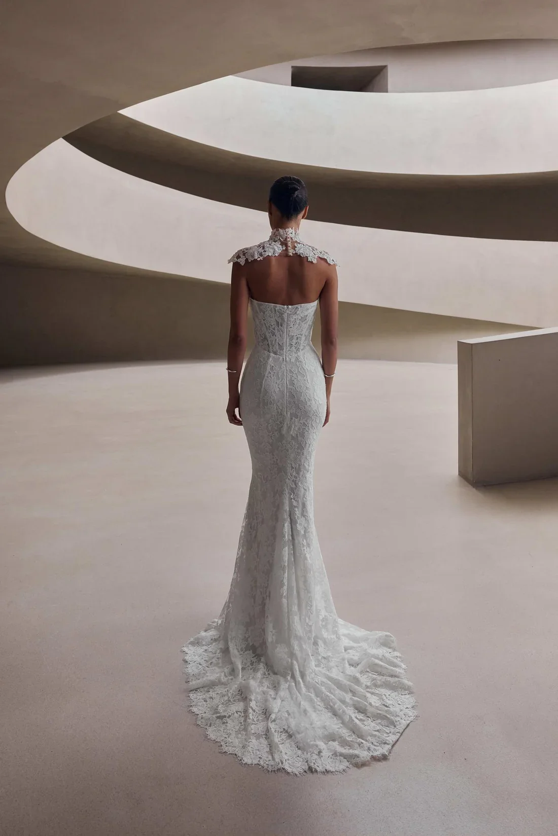 A woman in a strapless, lace wedding dress standing with her back to the camera in a modern, minimalist interior with curved architectural features.