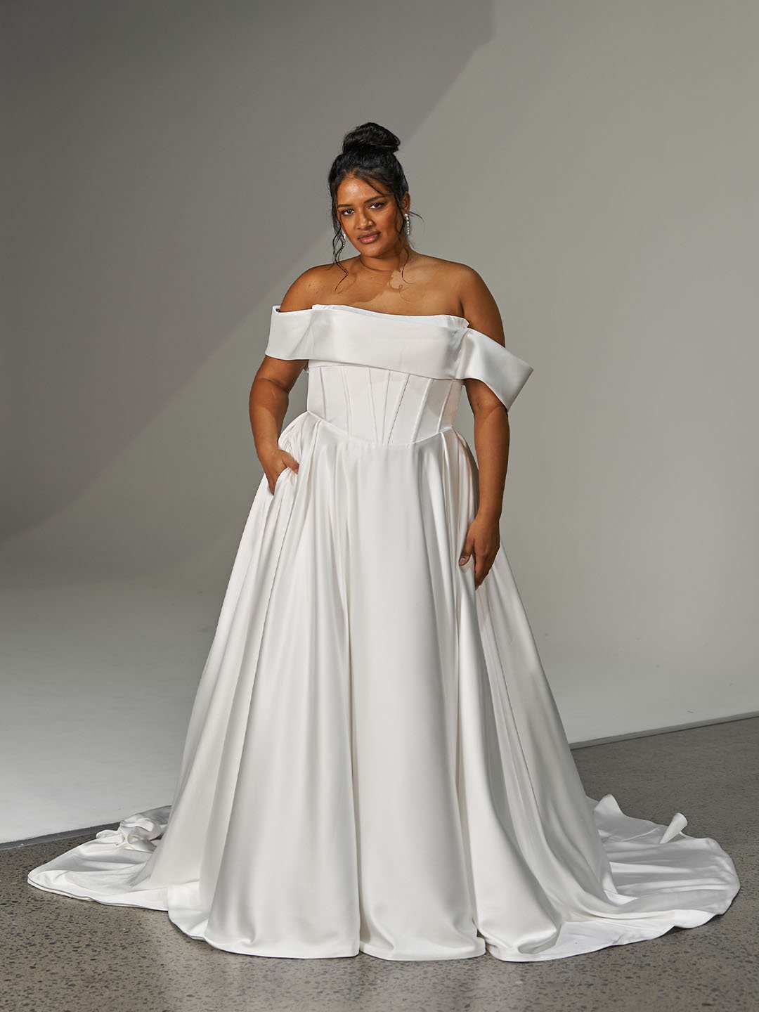 Woman wearing an off-the-shoulder white wedding dress standing against a studio background.