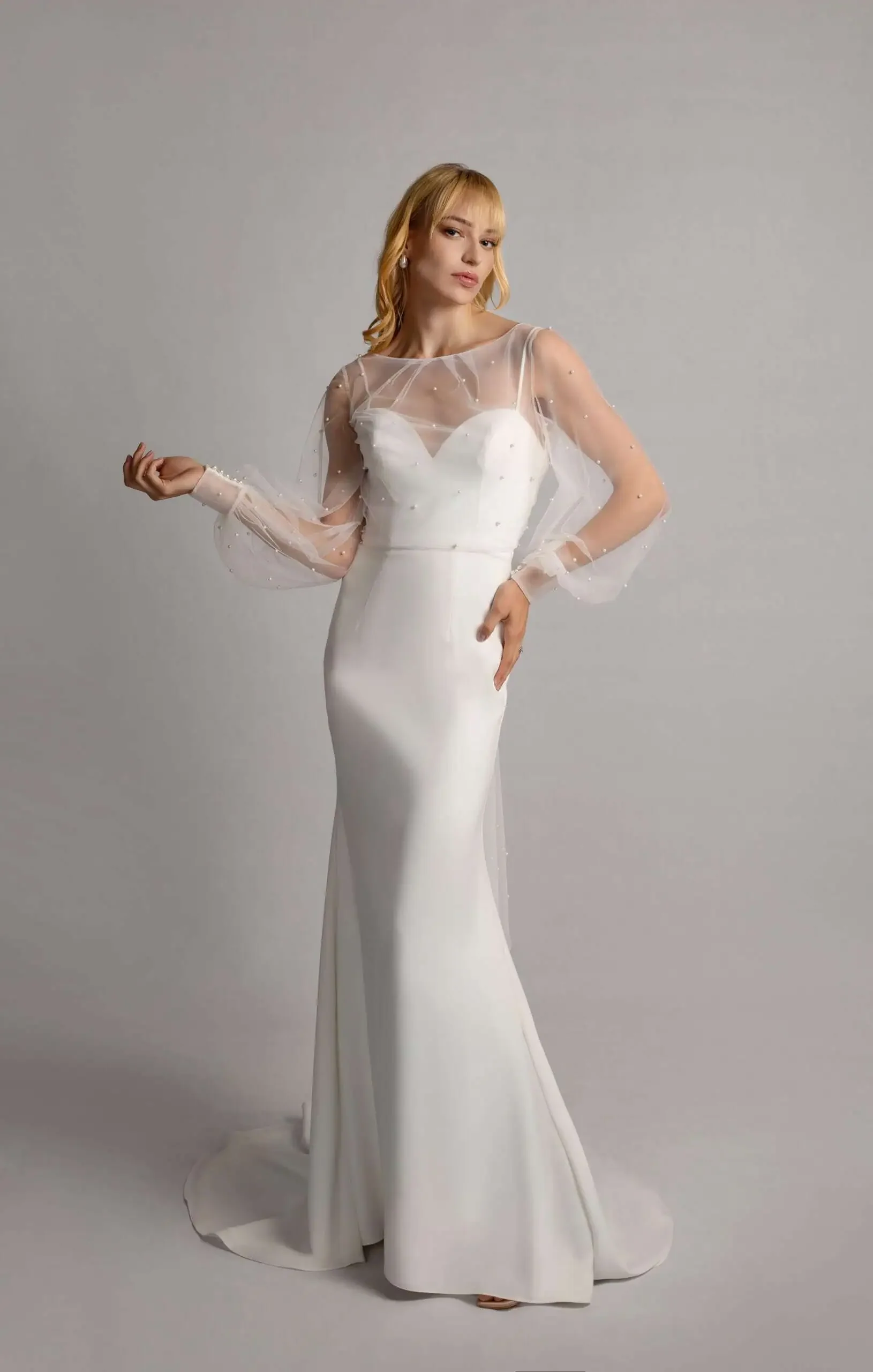 A woman in a fitted white wedding dress with sheer, dotted long sleeves and a sheer neckline, standing against a plain gray background.