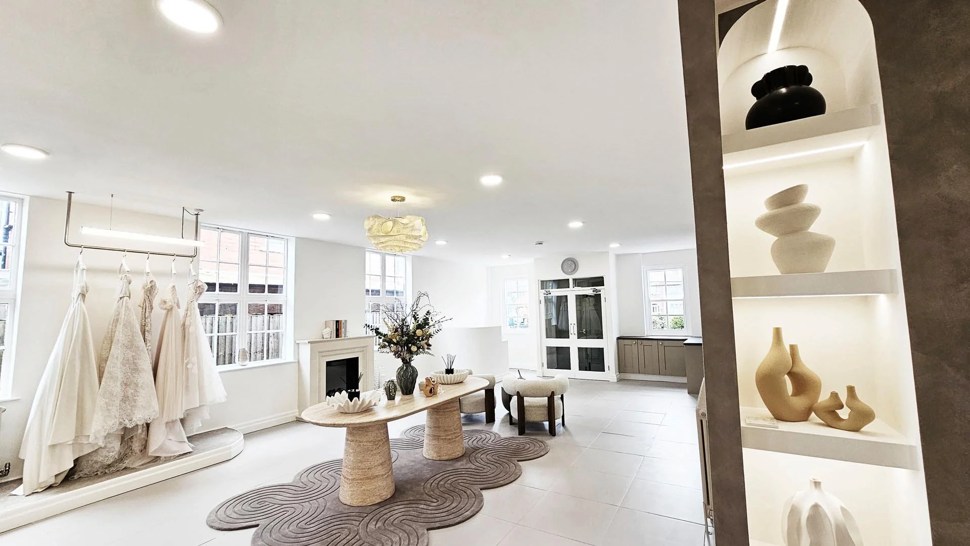 Bright, spacious interior with white walls, large windows, and modern decor. A display of white wedding dresses hangs on a rail, with a seating area featuring a round table, vases, and chairs. Decorative vases are on shelves to the right.