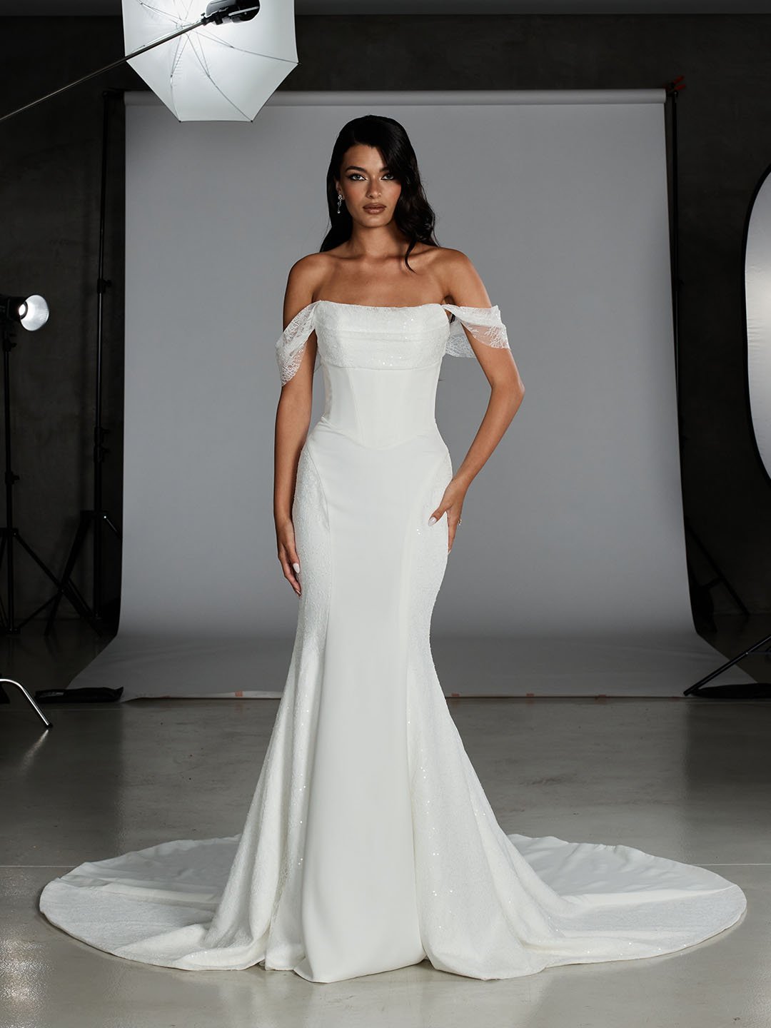 A woman wearing an off-the-shoulder white wedding gown with lace sleeves, standing on a white fabric train in a photography studio with a gray backdrop and studio lights.