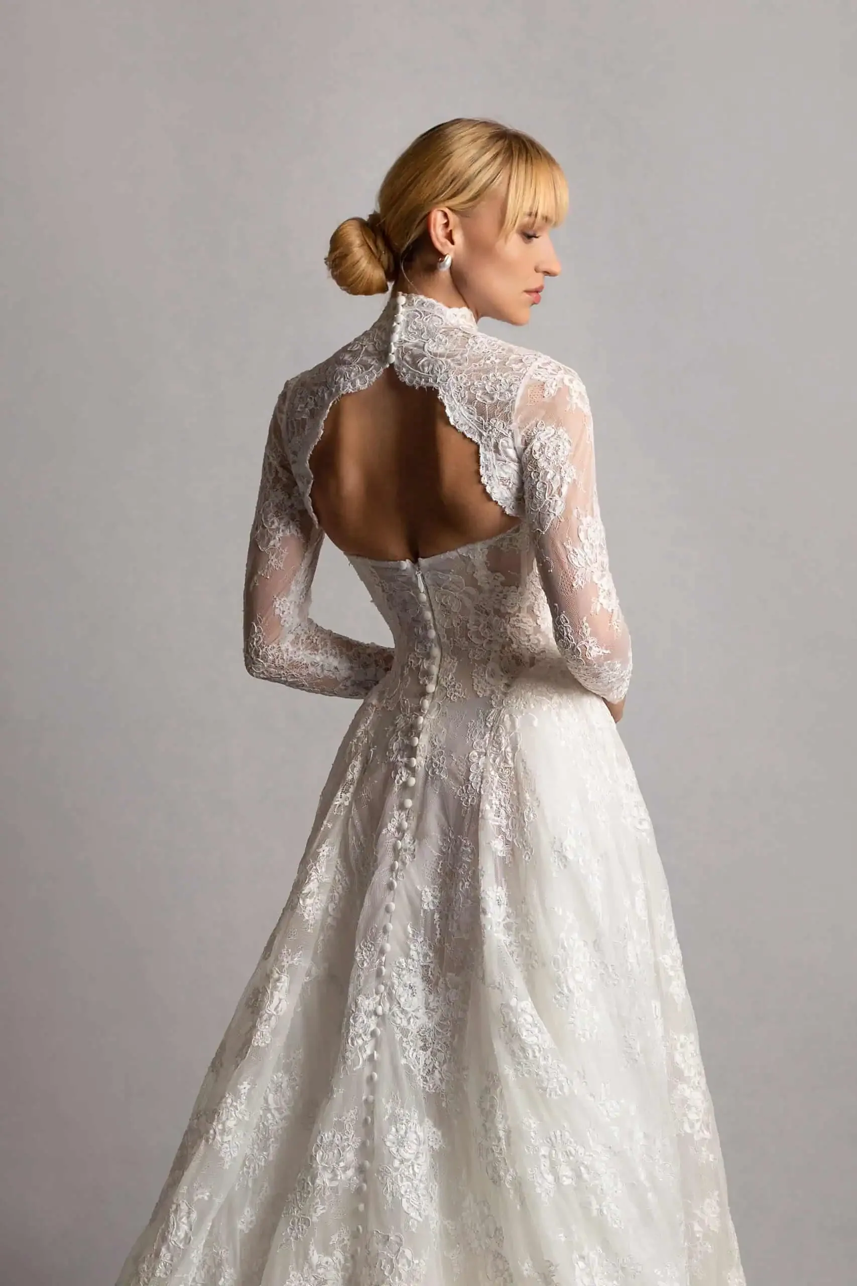 A woman with blonde hair styled in a low bun, wearing a white lace wedding dress with long sleeves and a high neck, standing with her back to the camera, looking to the side against a simple gray background.
