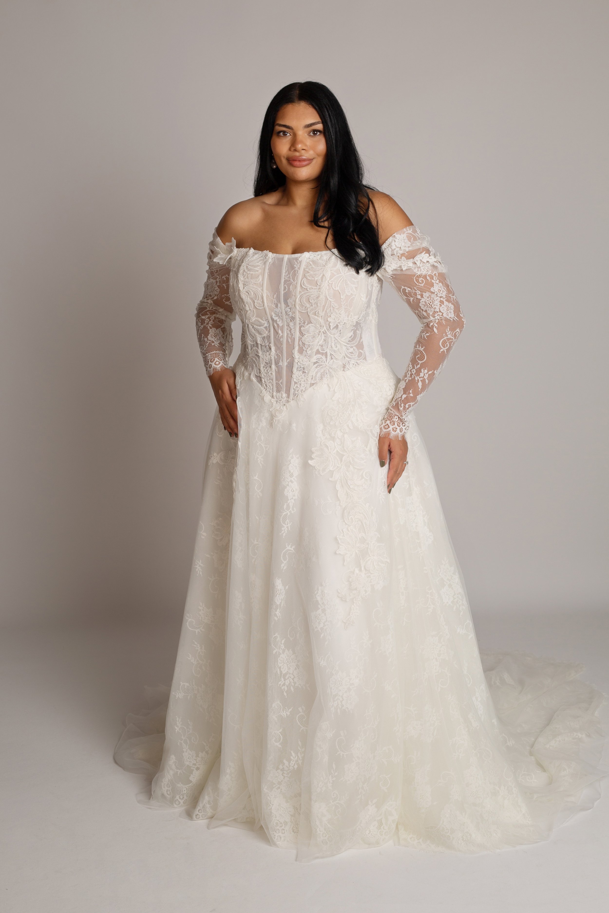 Woman in a white lace wedding dress standing against a plain background.