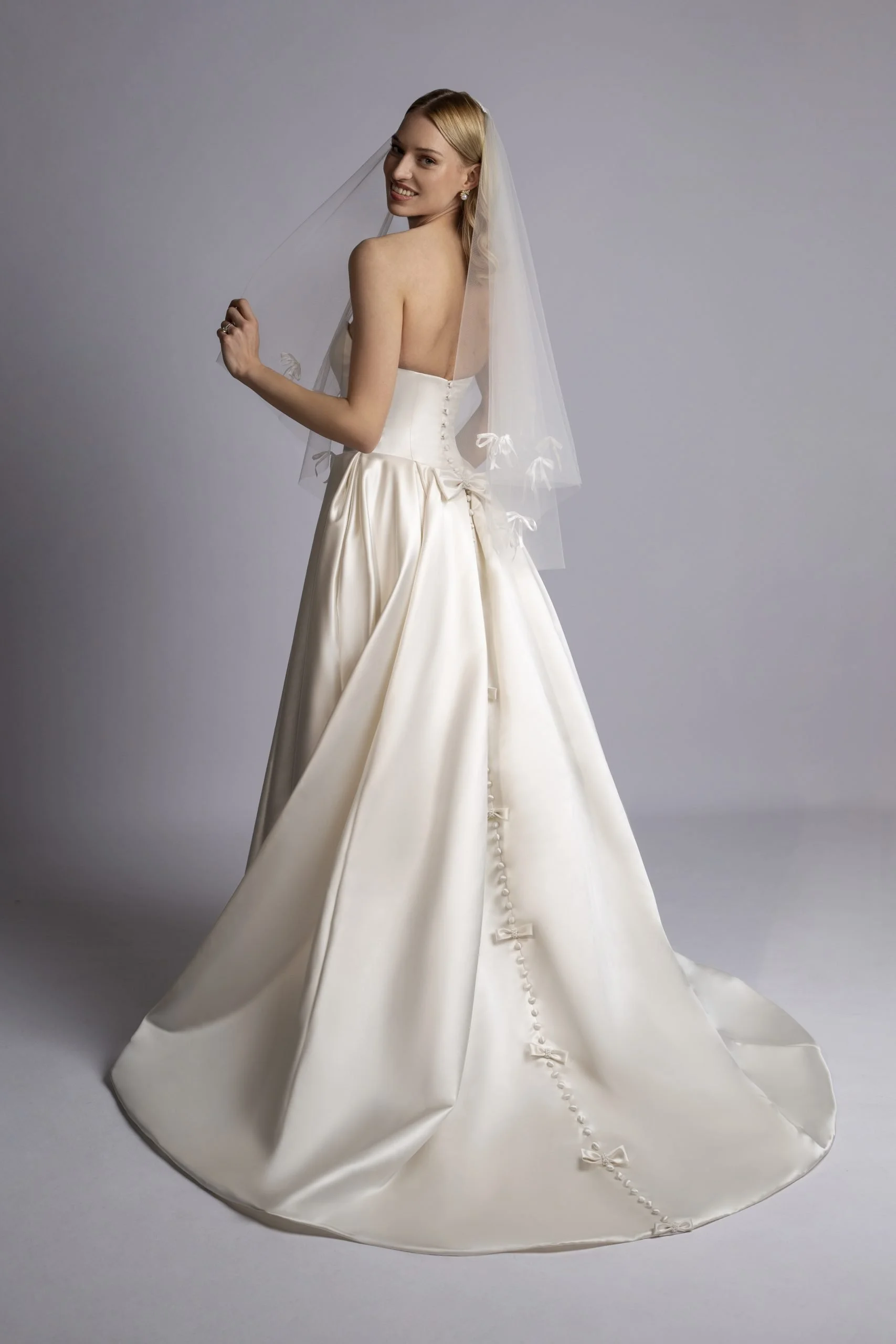 A woman in a wedding dress with a veil, smiling and looking over her shoulder.