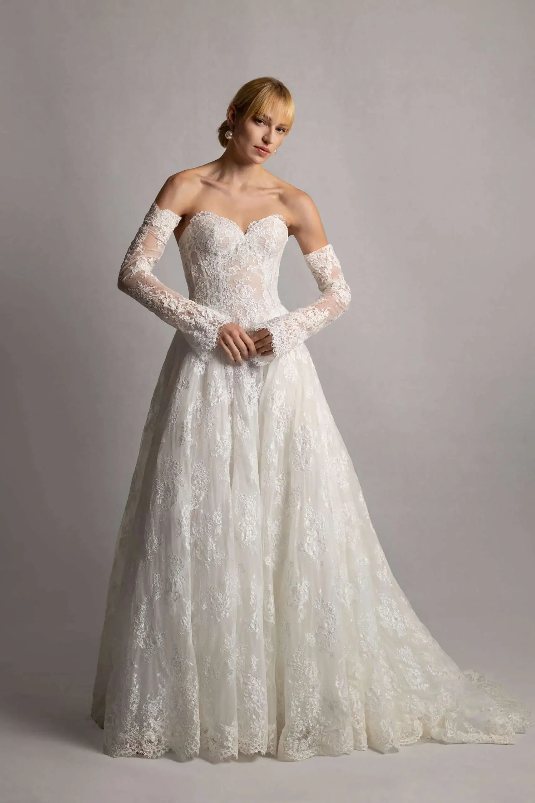 A woman in a strapless, long-sleeved white lace wedding dress standing against a plain gray background.