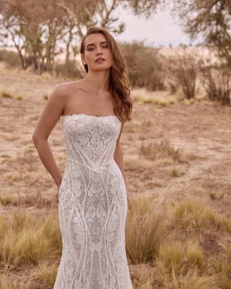 A woman stands outdoors in a desert landscape, wearing a strapless, white lace wedding dress with intricate patterns.