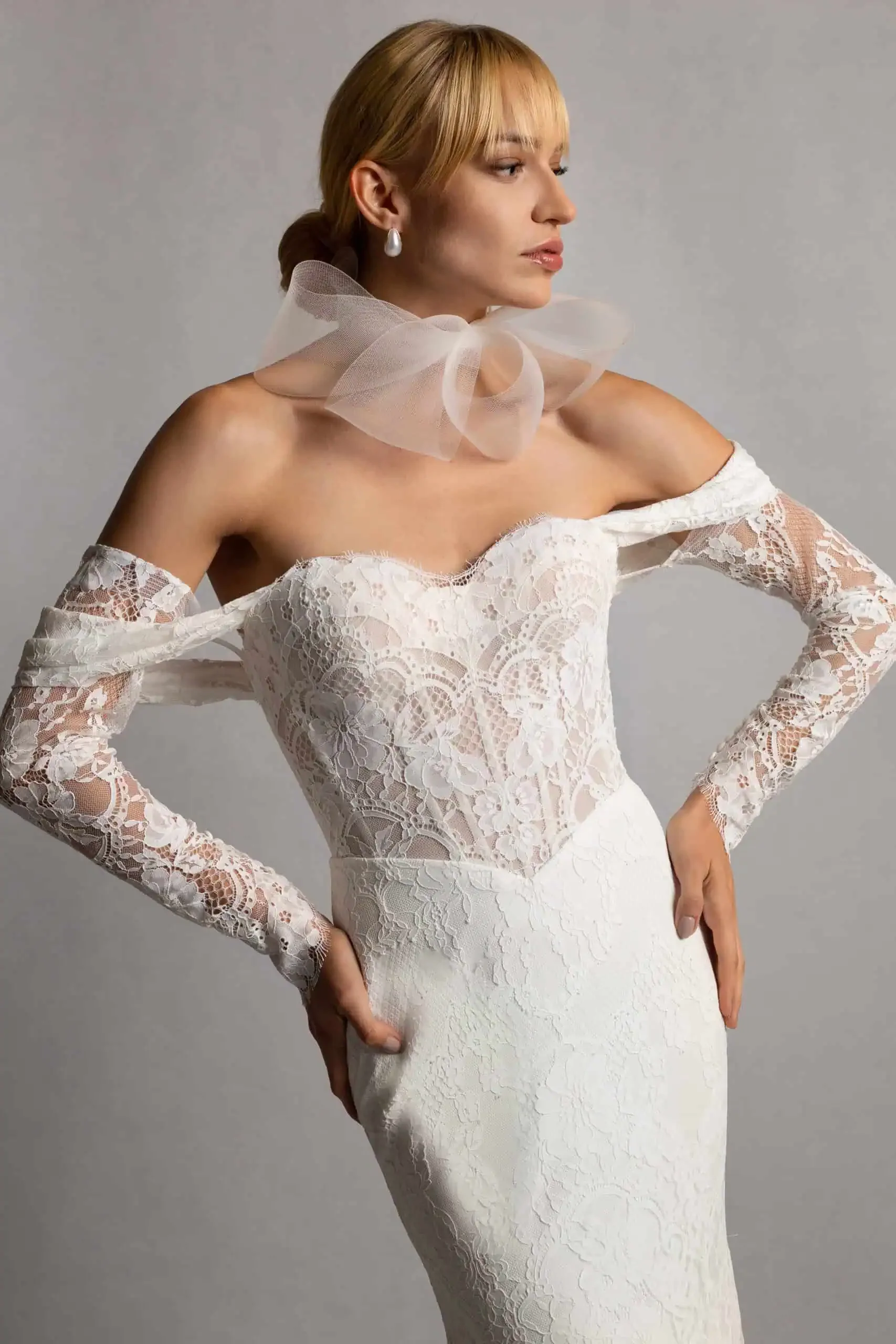 A woman wearing a white lace off-the-shoulder wedding dress with long lace sleeves, accessorized with pearl earrings and a sheer tulle bow around her neck, standing against a gray background.