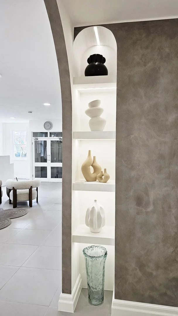 A built-in white display shelf with multi-tiered lighting, showcasing decorative vases and a black sculpture, situated in a modern interior space with gray walls and tiled flooring.