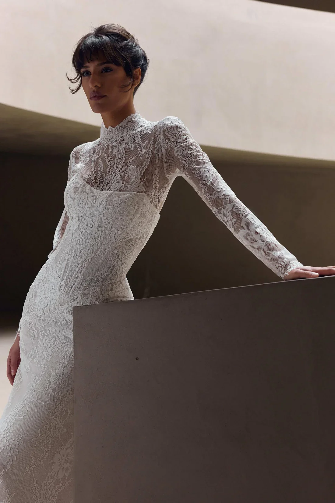 A woman in a white lace long-sleeve dress with a high neckline leaning on a gray railing in a modern architectural setting.