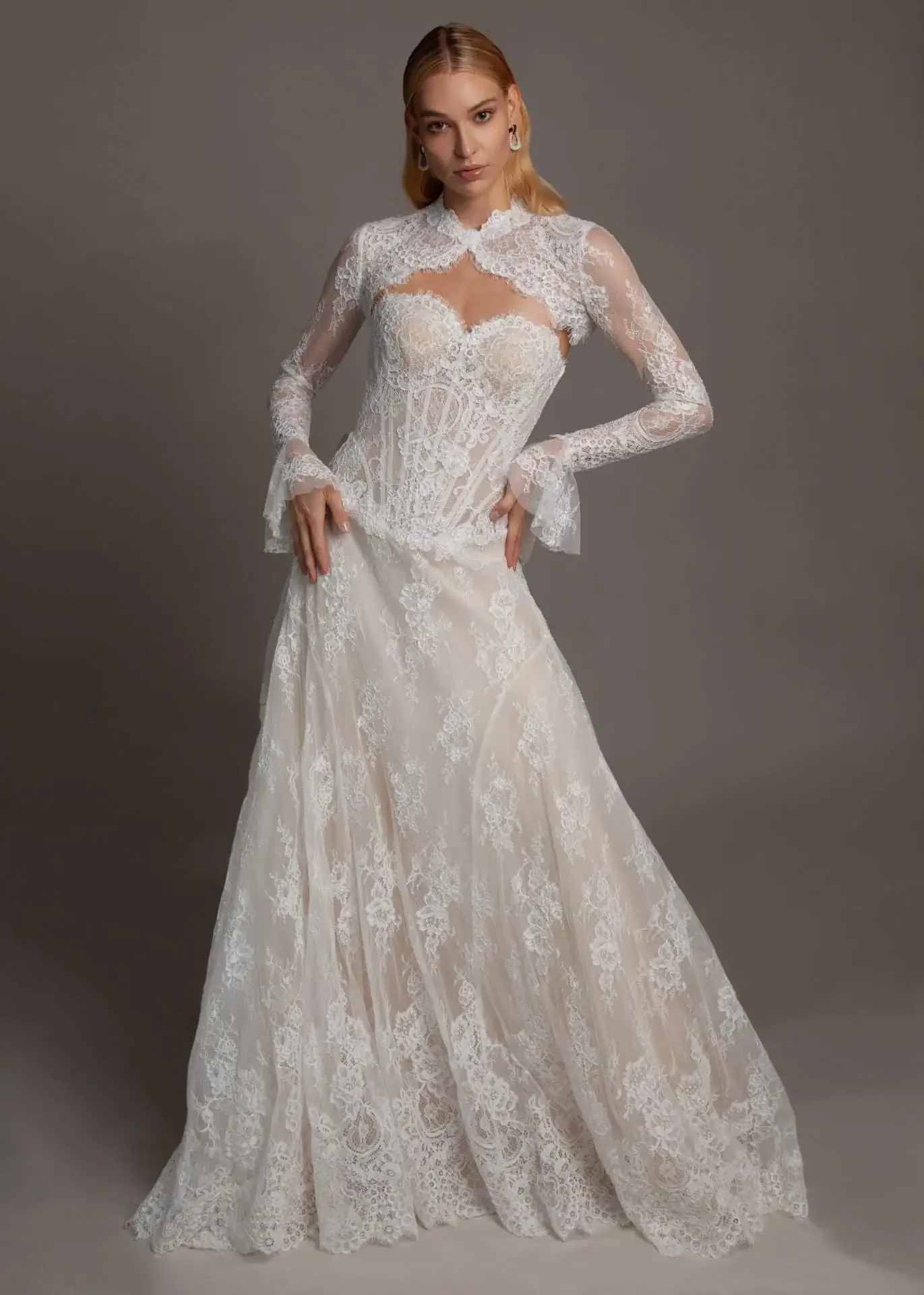 A woman in a white lace wedding dress with long sleeves and a high neck, standing against a plain gray background.
