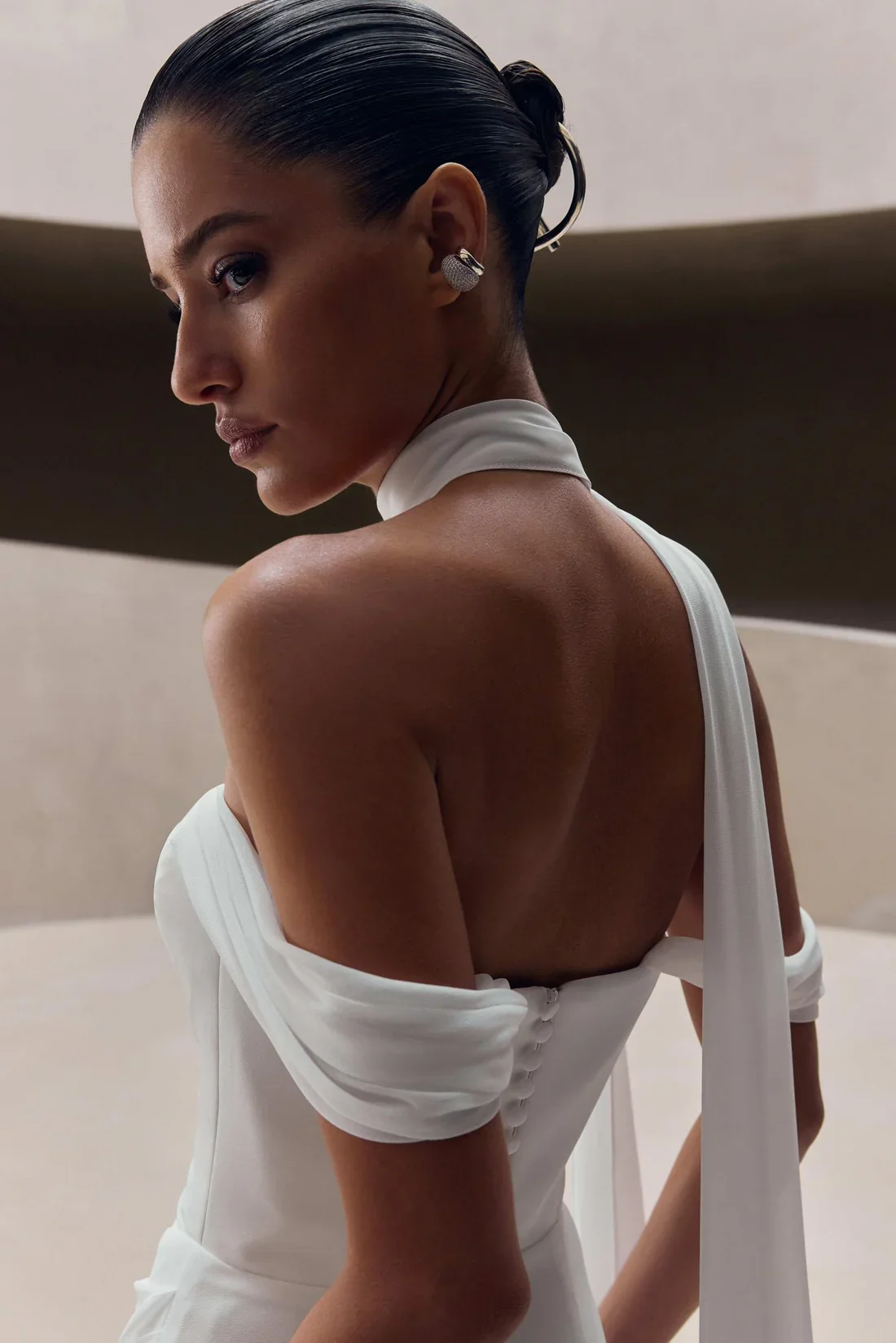 A woman with sleek, dark hair in a bun, wearing large earrings, posed in a white dress with an open back, sitting against a neutral background.