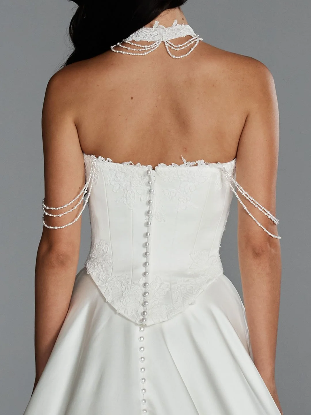 Back view of a woman wearing a strapless white wedding gown with pearl button details and pearl jewelry draped over her shoulders and neck.