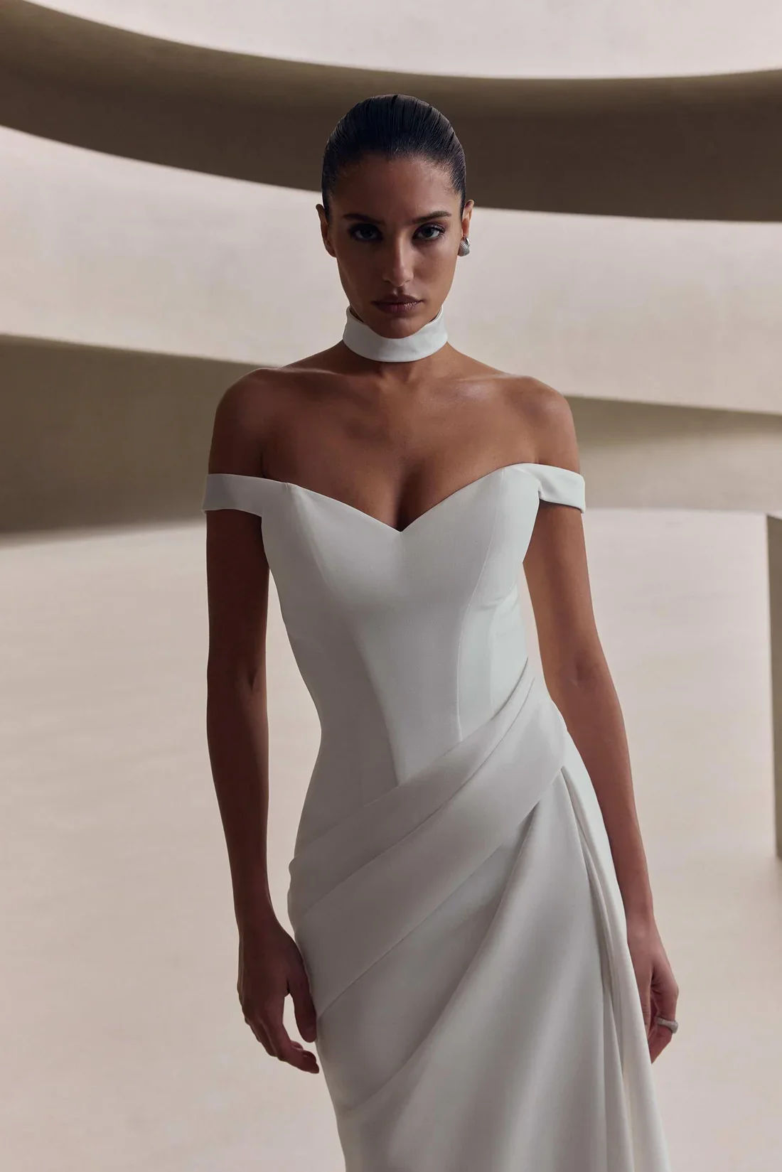 Woman in an off-the-shoulder white dress with a choker, standing in front of a modern, minimalist background.