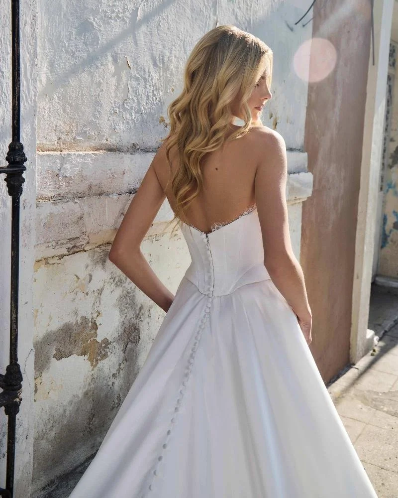 A woman in a white strapless wedding dress with wavy blonde hair, standing outdoors against a weathered white wall.