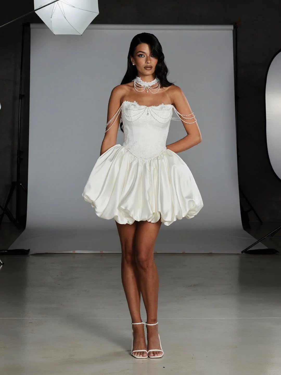 A woman posing in a studio wearing a white, off-the-shoulder, pearl-embellished dress with a puffed skirt, pearl jewelry, and high heels.
