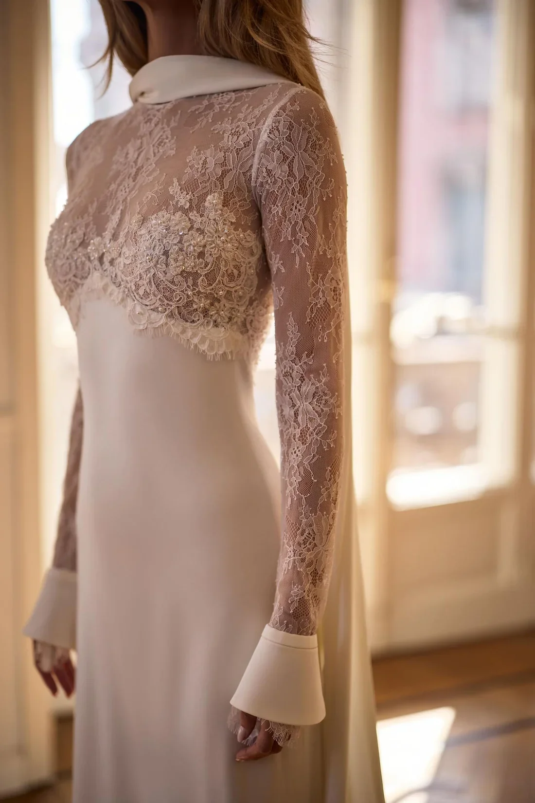 A woman wearing a vintage cream-colored dress with lace sleeves and a high collar, standing indoors near a window with sunlight streaming in.