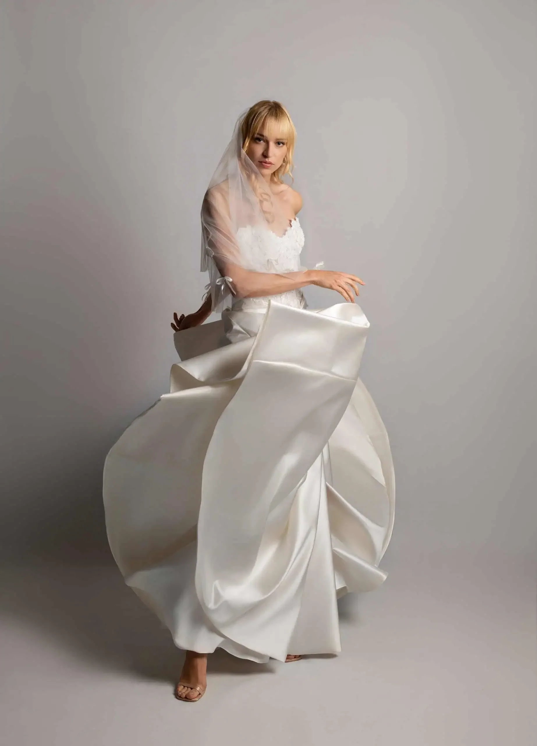 A woman in a white wedding dress with a sheer veil stands against a light gray background.