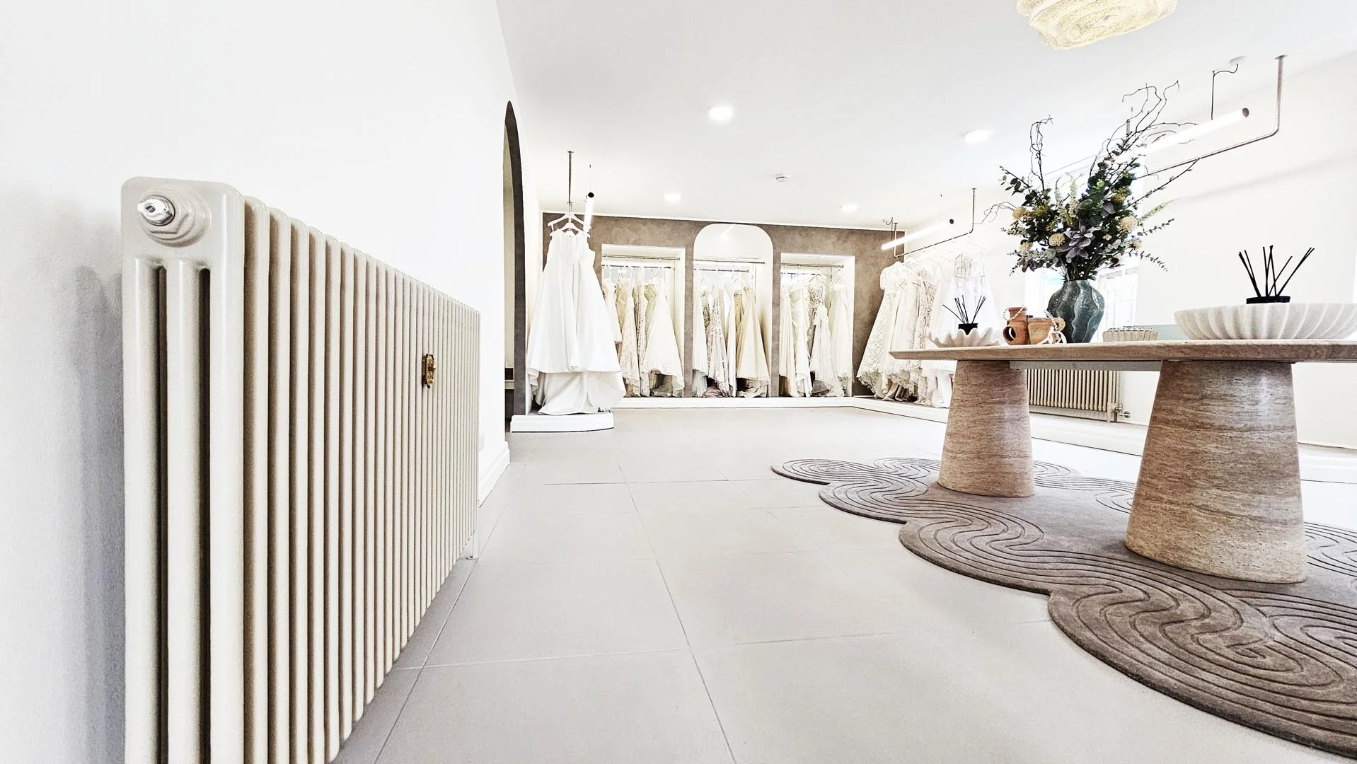 A bridal boutique with wedding dresses hanging on racks near the windows, a round display table with a vase of flowers, decorative bowls, and reed diffusers, and a cozy, neutral-toned rug.