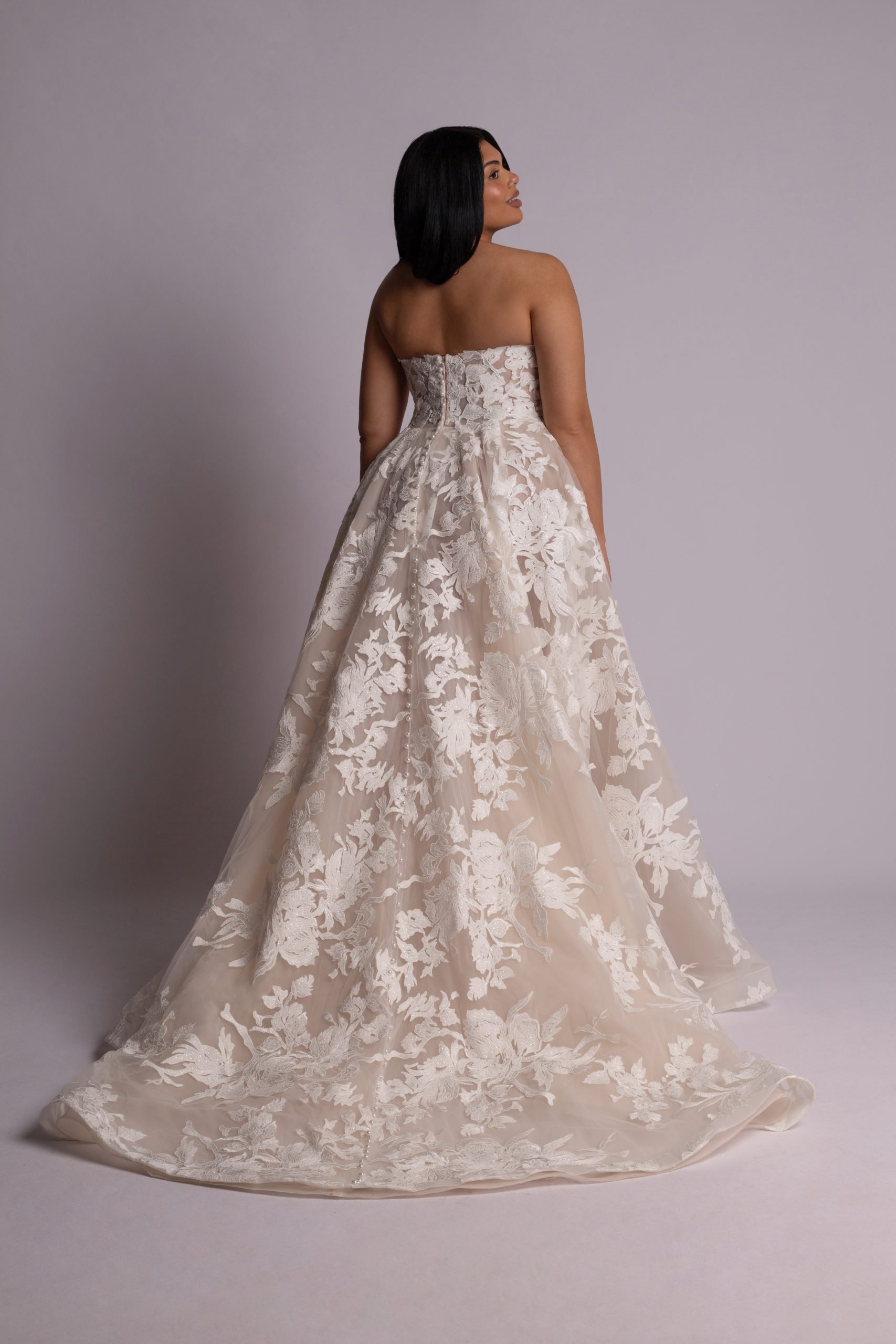 A woman wearing a strapless wedding gown with lace and floral embroidery, standing against a plain background, with her back to the camera and head turned to the side.