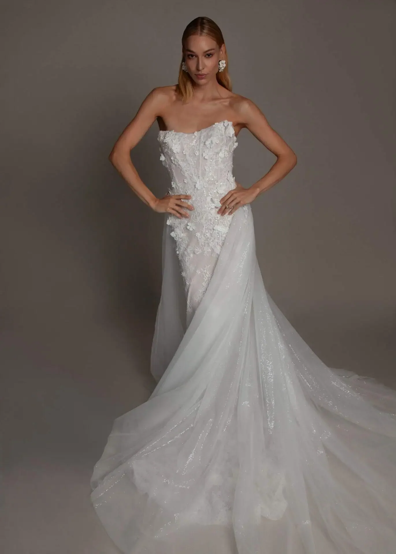A woman with red hair wearing a strapless white wedding dress with floral lace appliqué and a train.