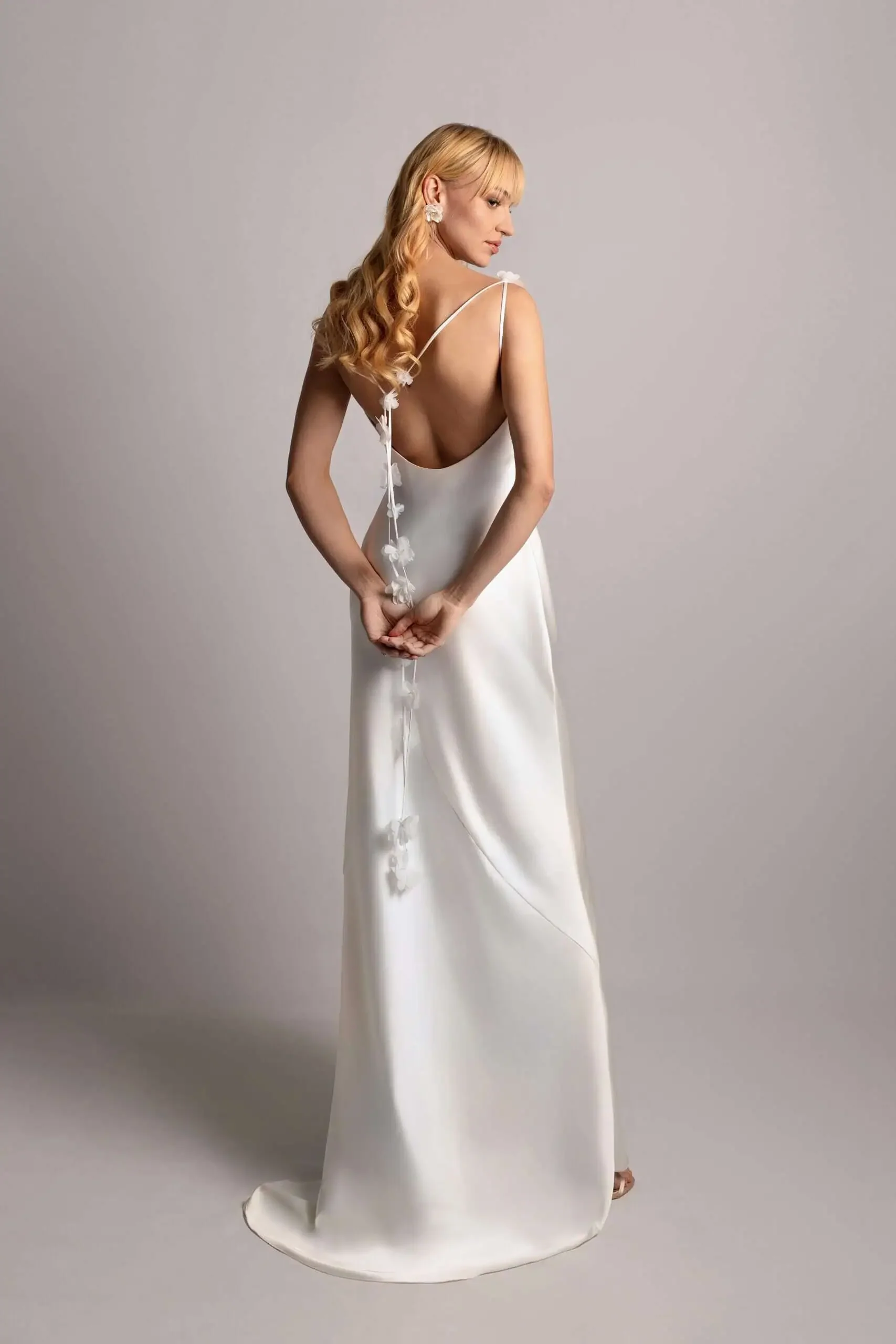 A woman with blonde hair in loose waves wearing a backless white satin dress with floral embellishments, standing against a plain gray background.
