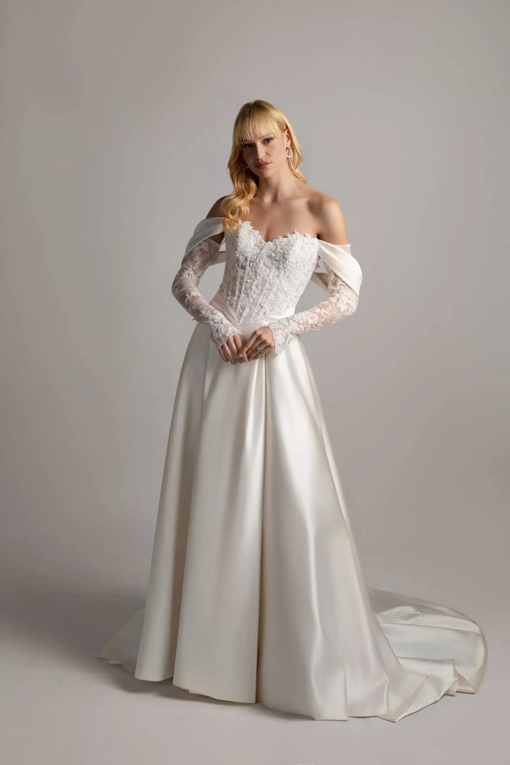 A woman wearing an off-shoulder white wedding dress with lace sleeves and an embroidered bodice, standing against a plain background.