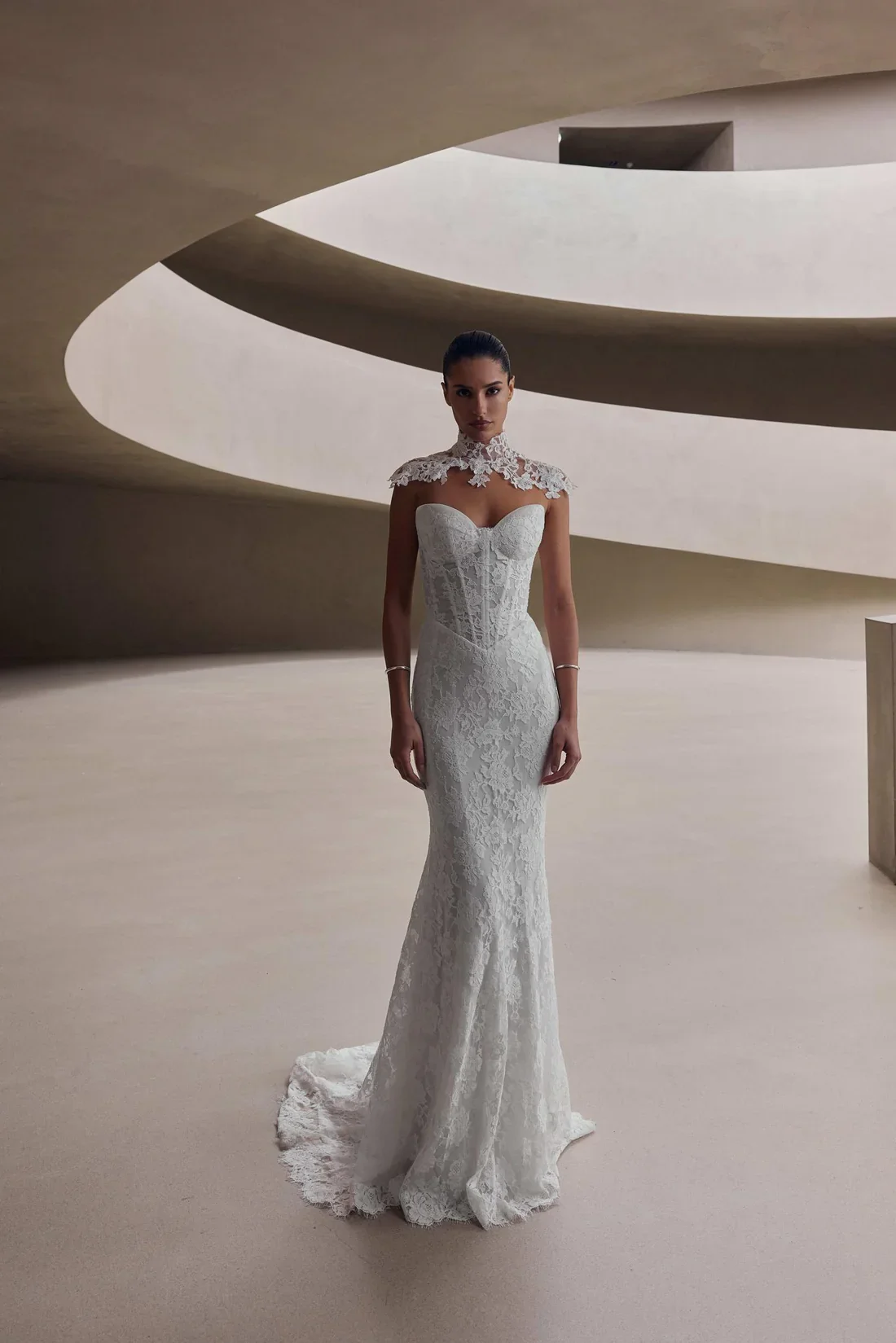 A woman in a white lace wedding dress standing in a modern, minimalist interior with curved architectural features.