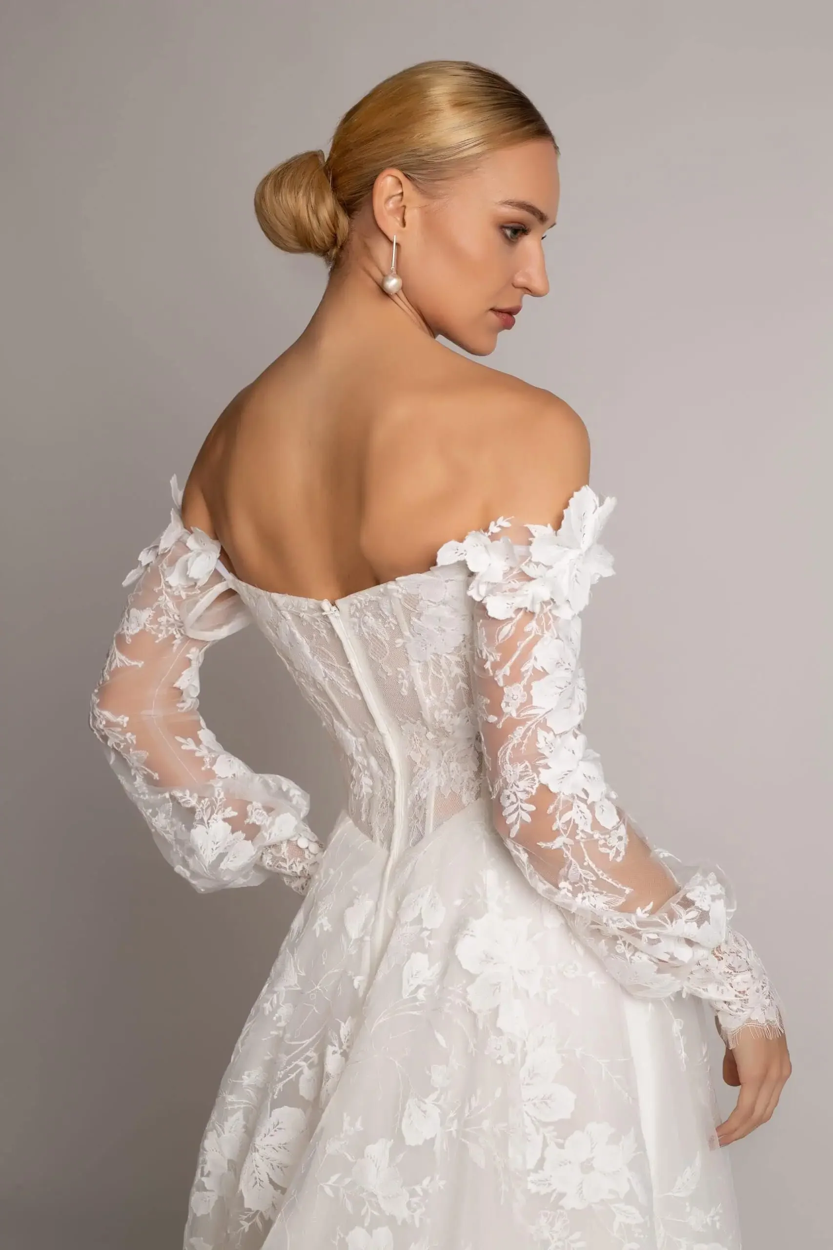 A woman in a white wedding dress with floral lace details, off-the-shoulder sleeves, and a fitted bodice, looking over her shoulder.