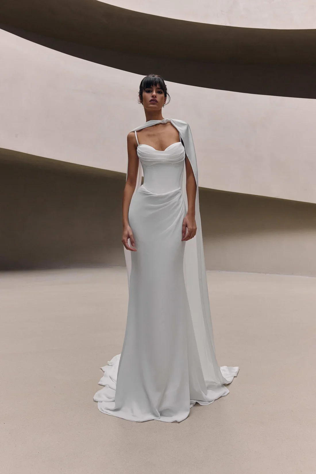 A woman in a long white bridal gown with a draped bodice and a flowing train, standing in an art gallery with curved beige walls.