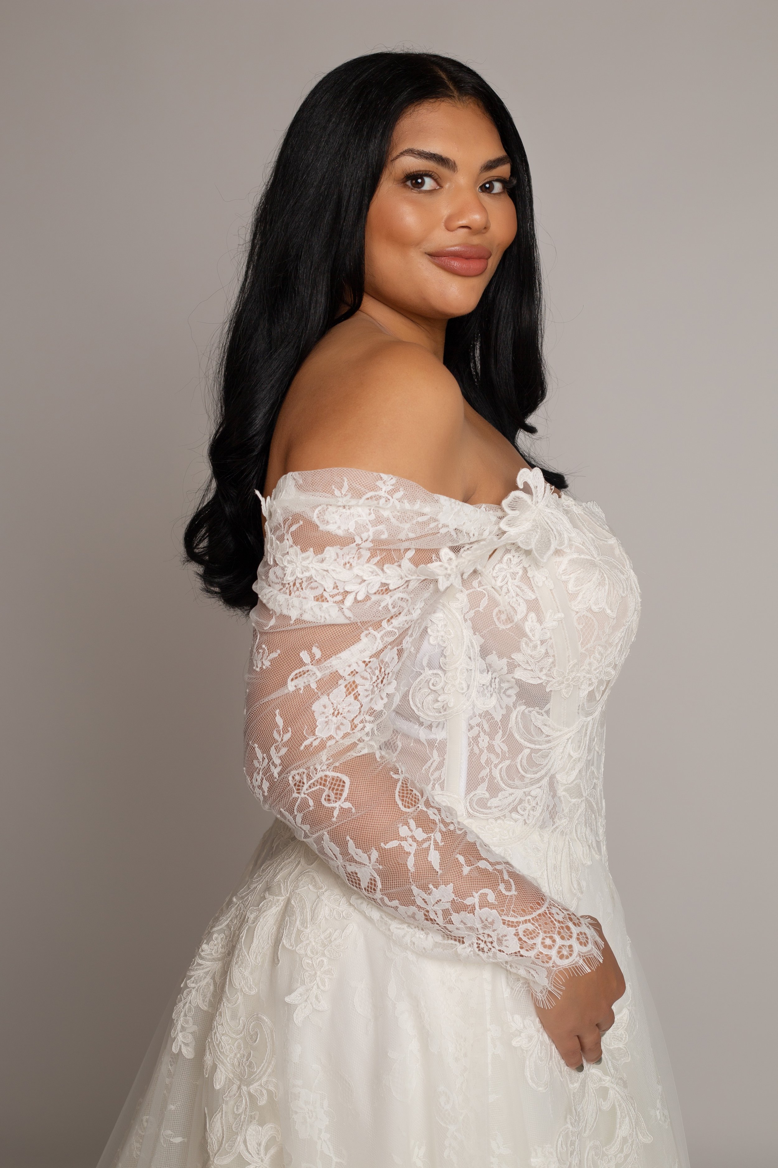 A woman with long dark hair wearing a white lace off-shoulder dress, posing against a plain light gray background.