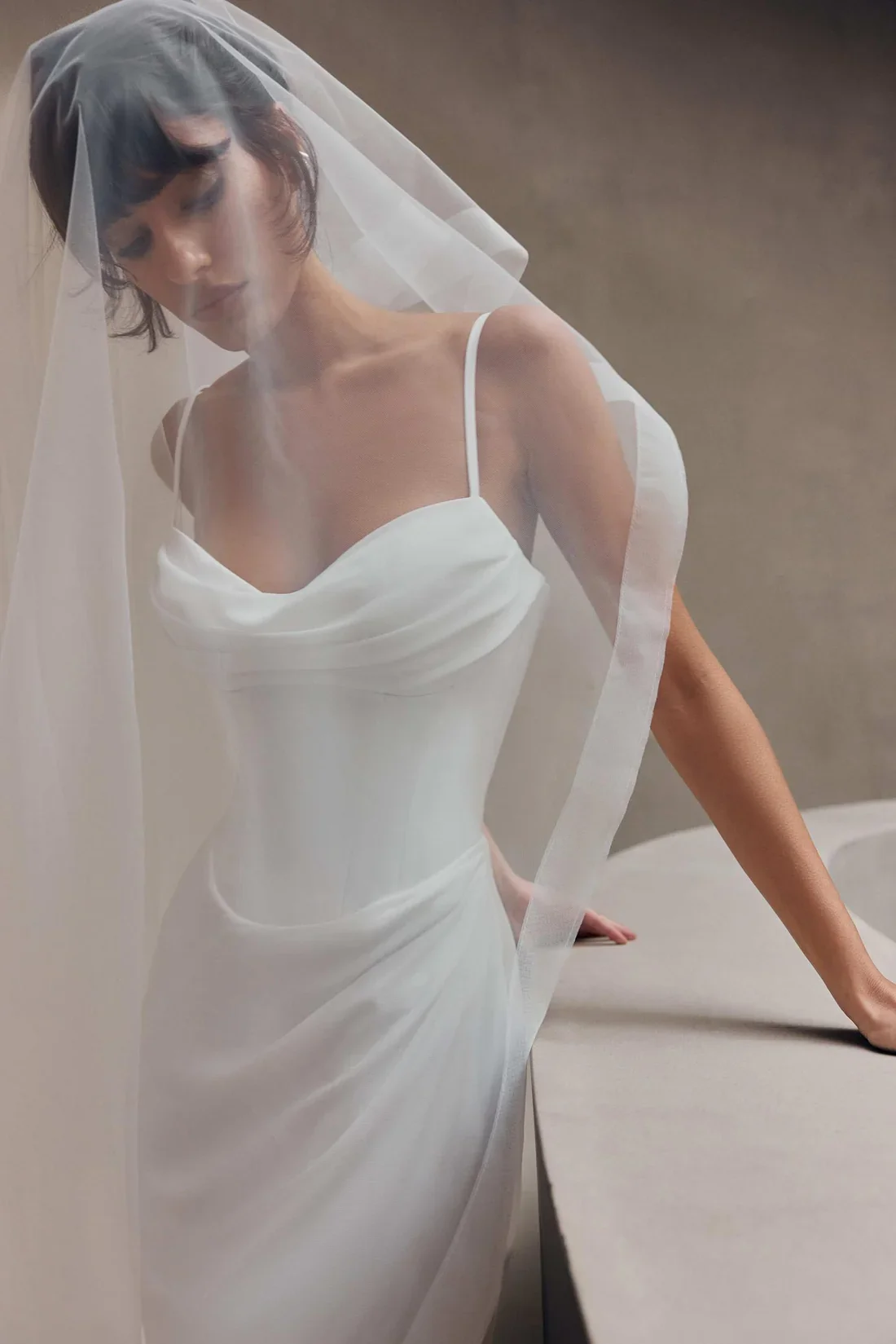 Woman in a white dress and veil leaning on a counter with a neutral background.