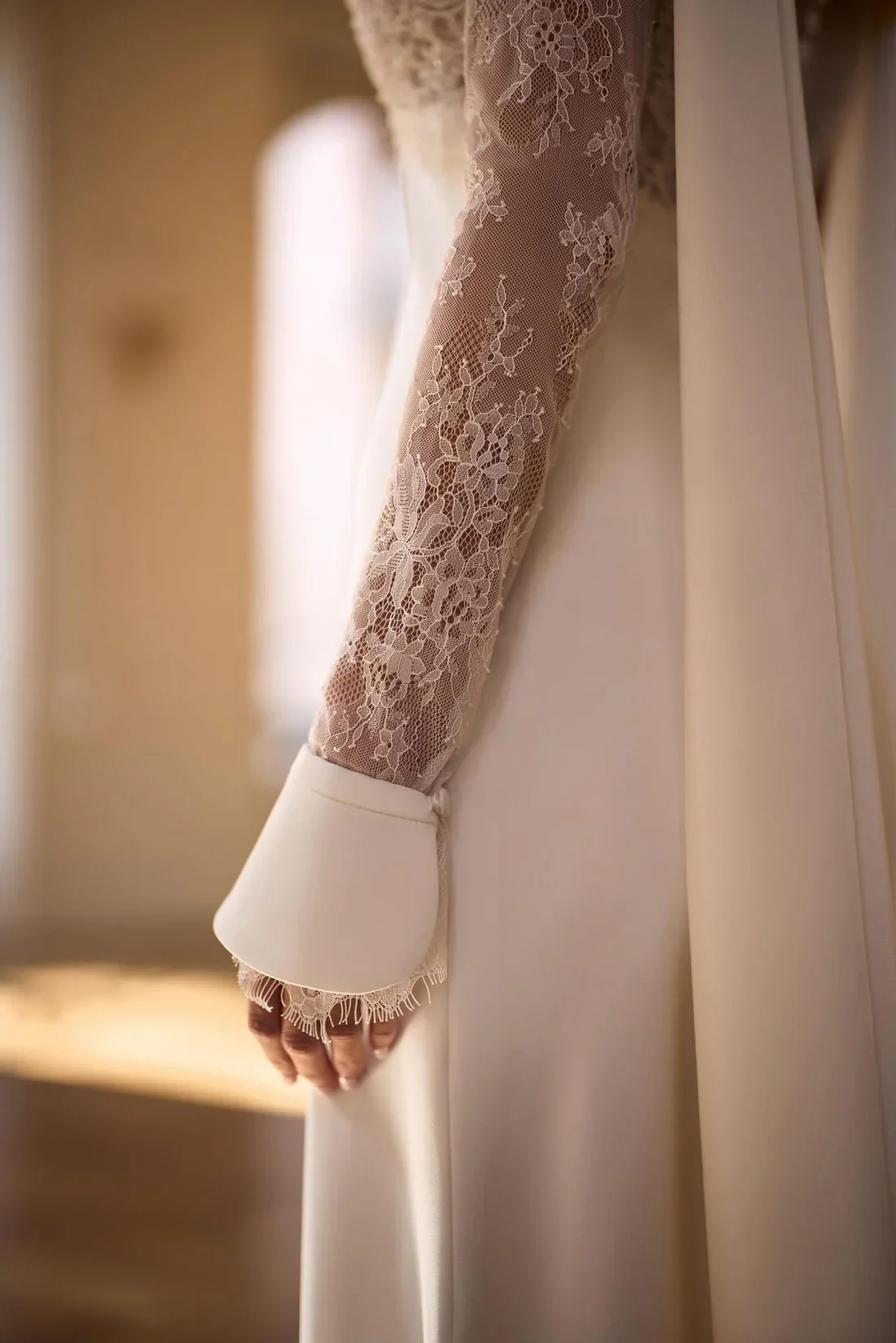 Close-up of a lace-covered sleeve of an elegant white garment, possibly a wedding dress, with a white cuff and delicate fringe detailing at the wrist.