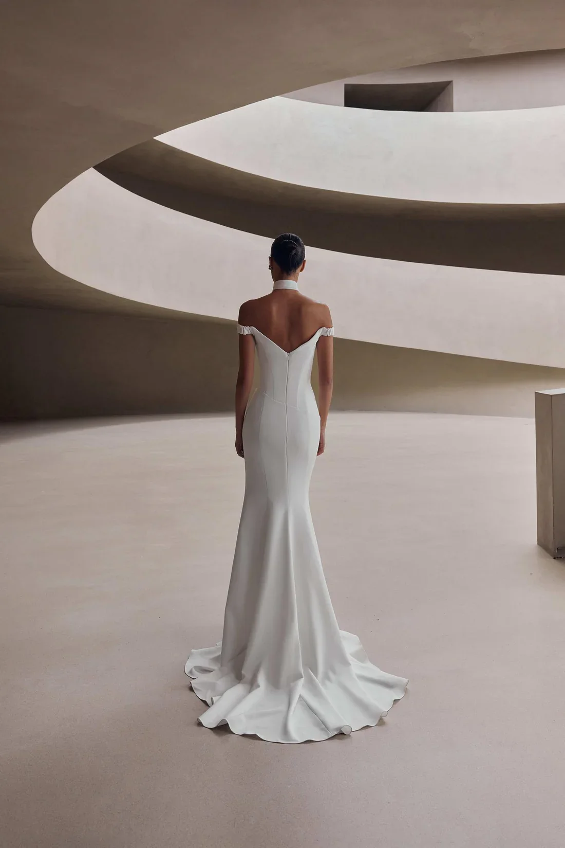 A woman in a white, elegant, off-shoulder wedding dress walking in a modern, minimalist architectural space with curved ceilings.
