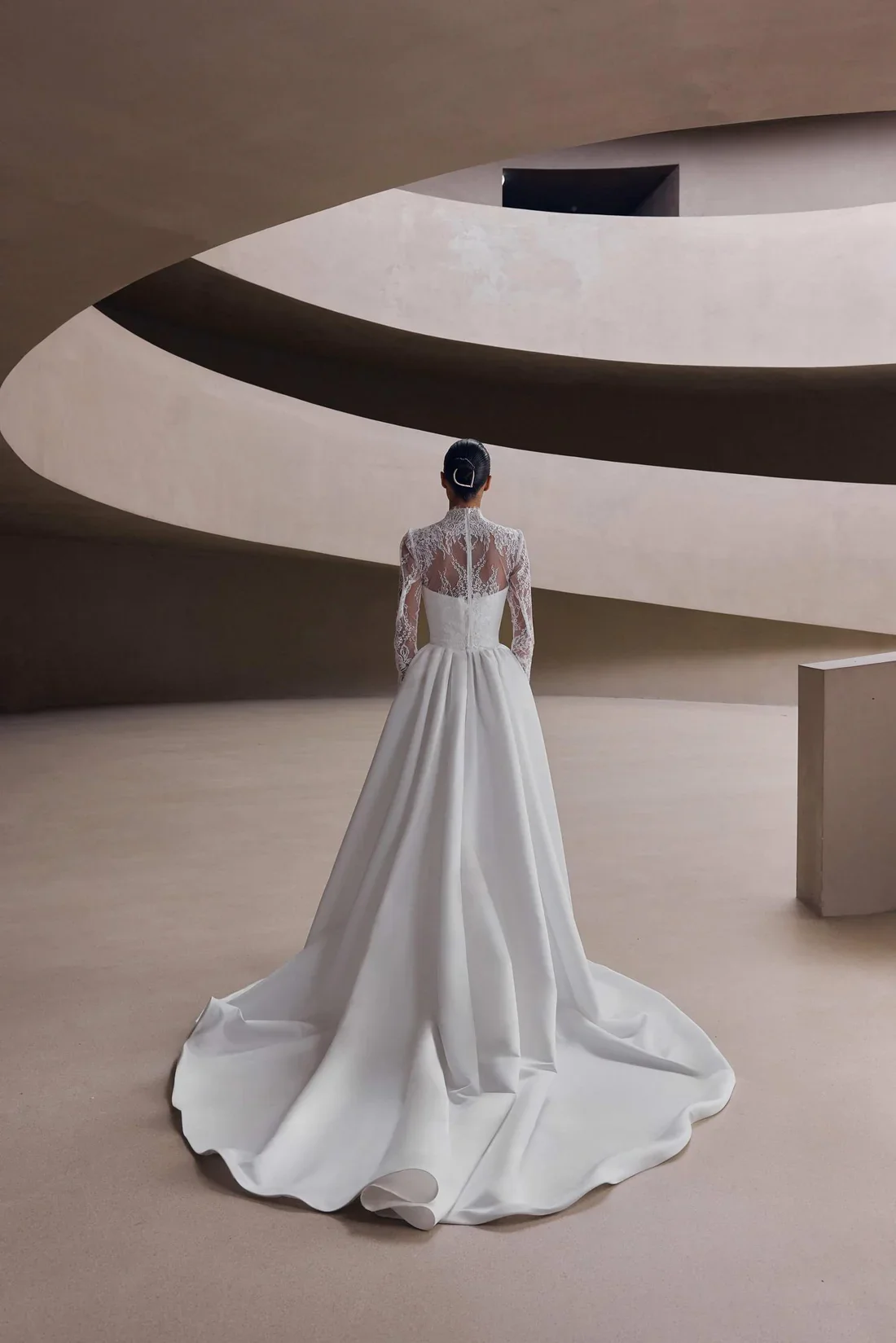A woman in a white wedding dress with lace top and long sleeves stands with her back to the camera in an architecturally modern, curved, minimalist interior.