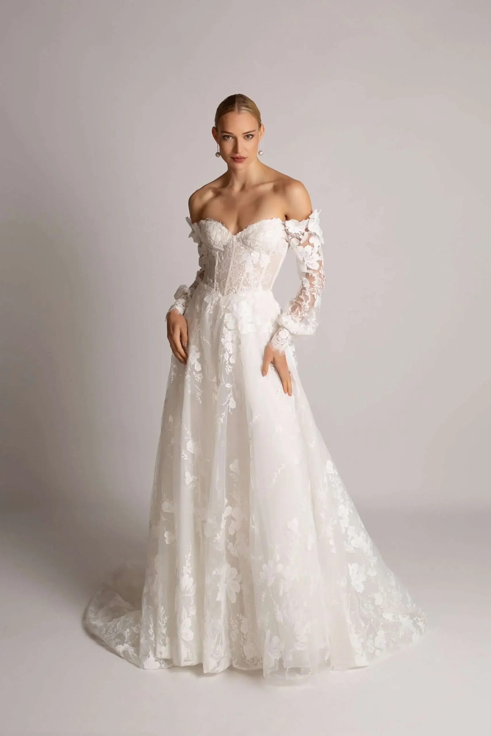 A woman wearing a white off-the-shoulder wedding dress with lace details, standing against a plain background.