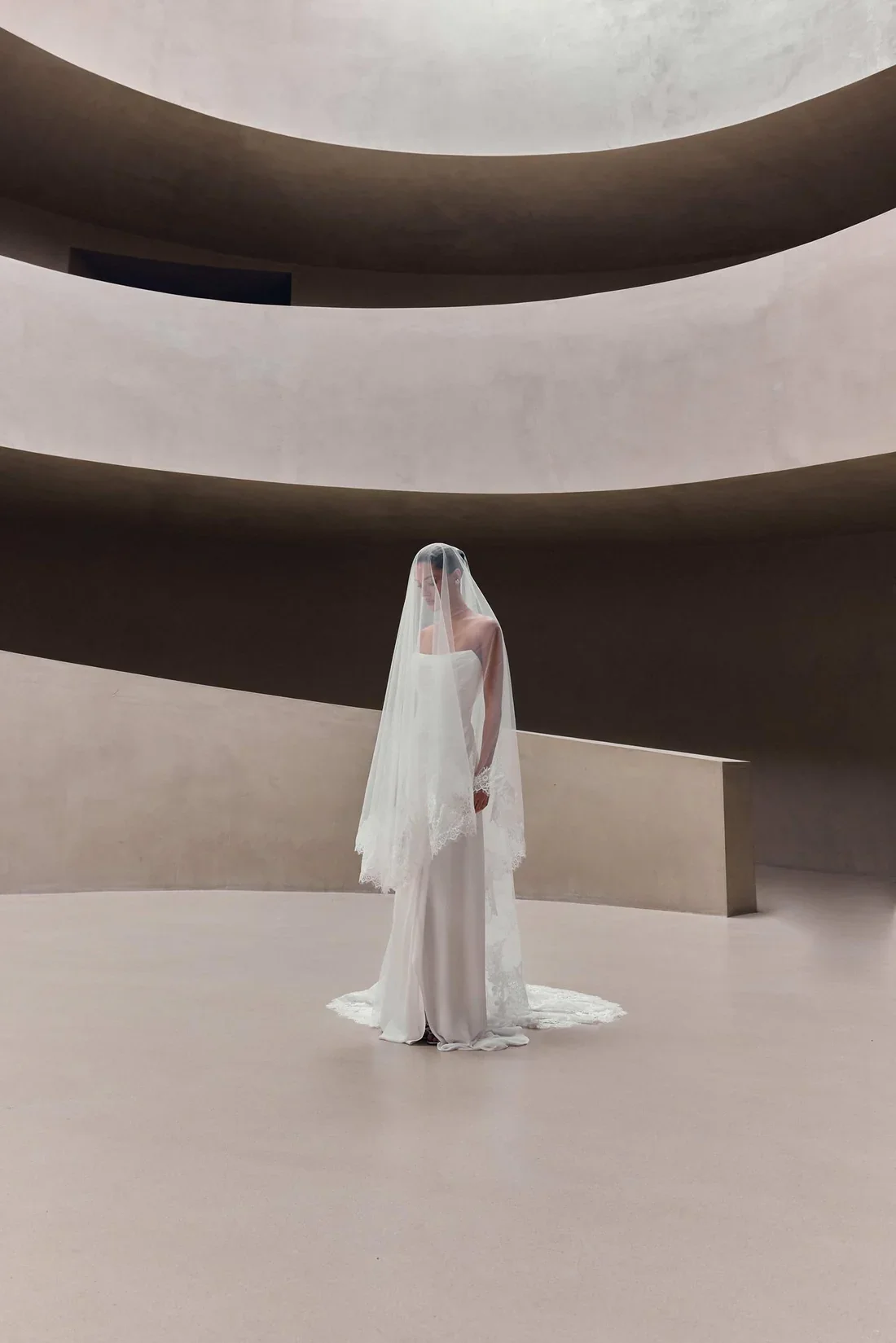 A bride in a wedding dress and veil standing alone in a modern, minimalist architectural space with curved, layered walls.