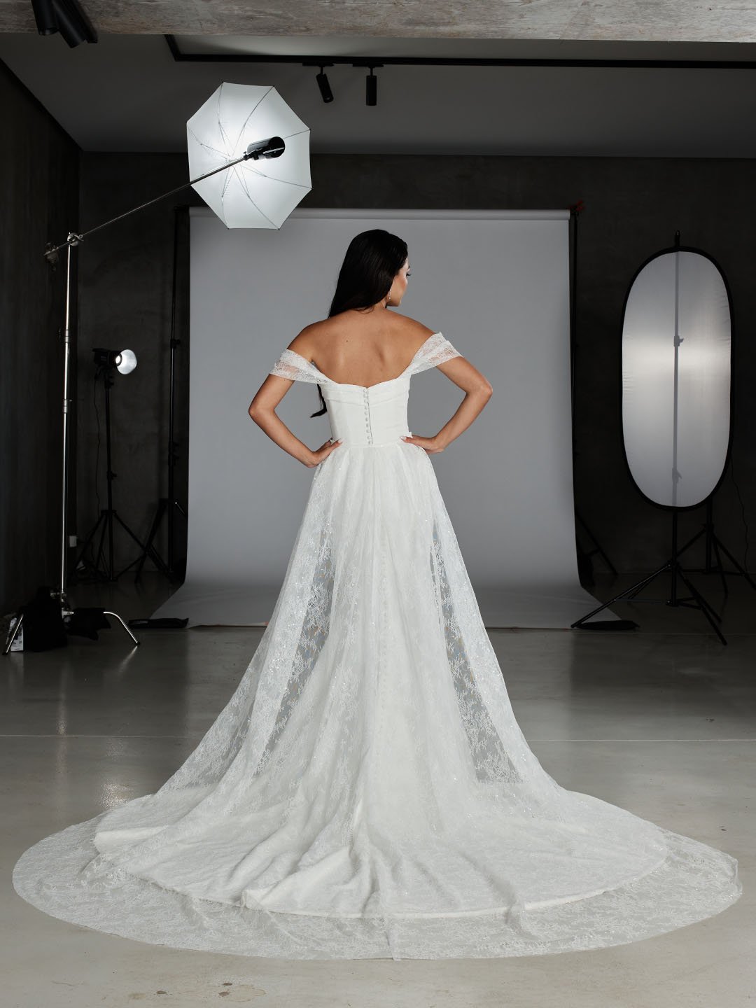 A woman in a white wedding dress with an off-shoulder lace design stands in a photography studio, facing away. The dress has a long train spread on the floor, and the studio has a backdrop with lighting equipment and reflectors.