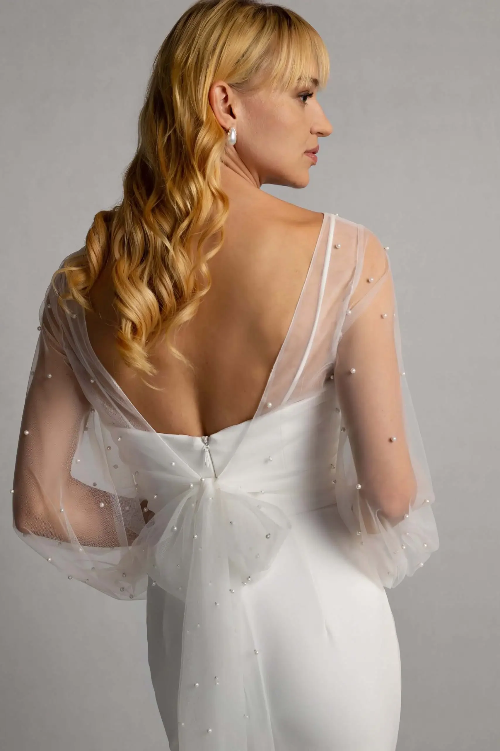 A woman in a white dress with sheer, beaded sleeves, facing sideways with her back turned, showing her profile. She has long, wavy blonde hair and is wearing pearl earrings.