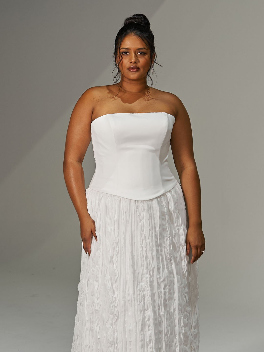 A woman standing in front of a neutral background, wearing a strapless white dress with a fitted bodice and a pleated, floor-length skirt.