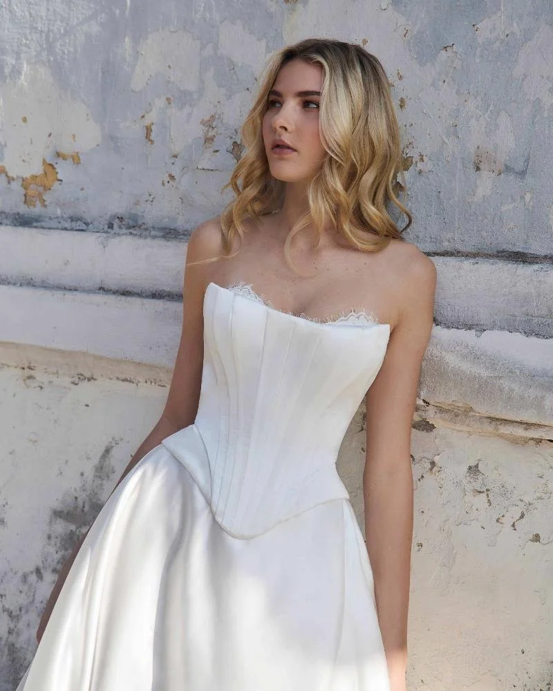 A young woman with blonde hair wearing a strapless white satin dress with a fitted bodice and a full skirt, standing against a textured stone wall.