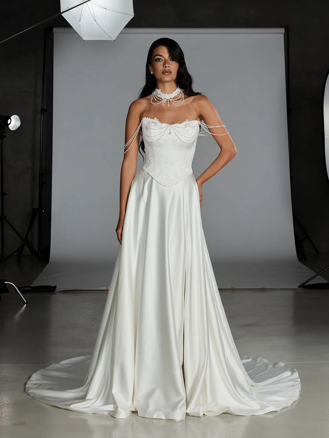A woman wearing a wedding dress with a strapless lace bodice and flowing satin skirt, adorned with pearl and chain jewelry, standing in a photography studio with a gray backdrop.