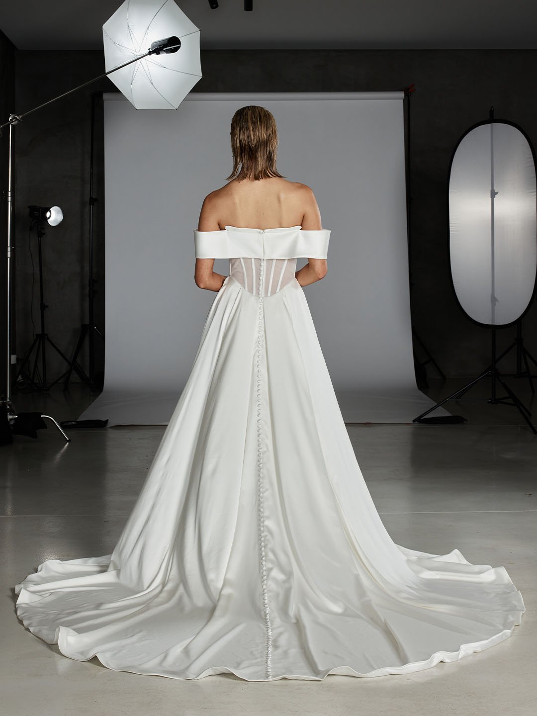 Back view of a woman in a white wedding gown standing in a photography studio with a white backdrop and studio lights.