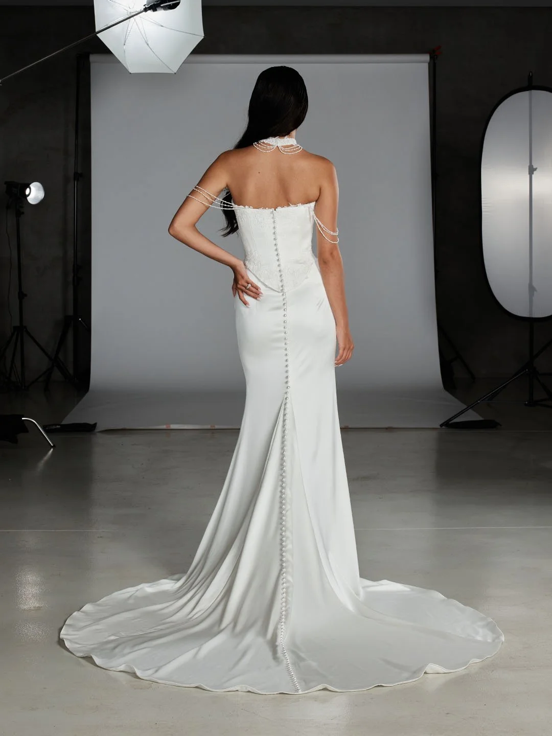 Woman in a strapless satin wedding dress with pearl button closure running down the back, standing in a photography studio.