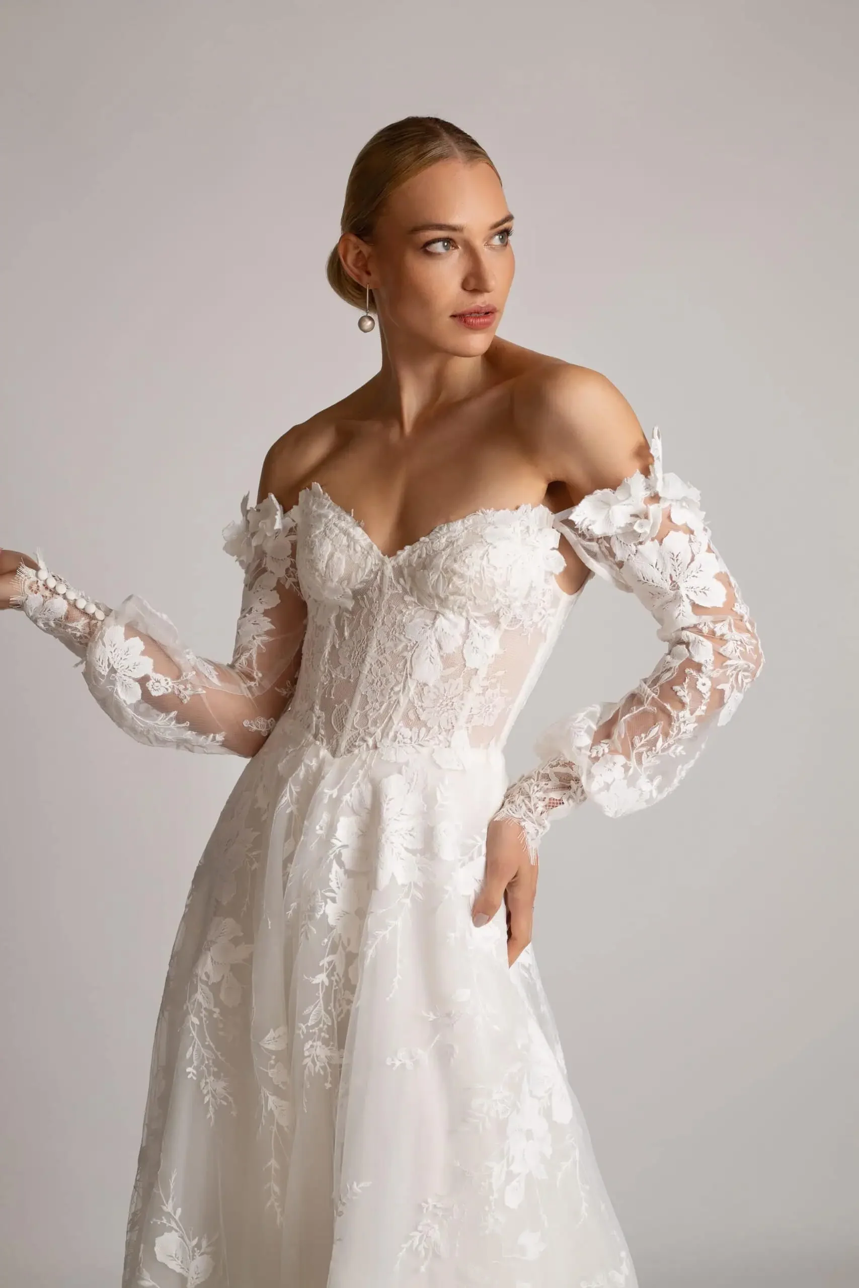 A woman in a white lace wedding dress with off-shoulder long sleeves and floral details.