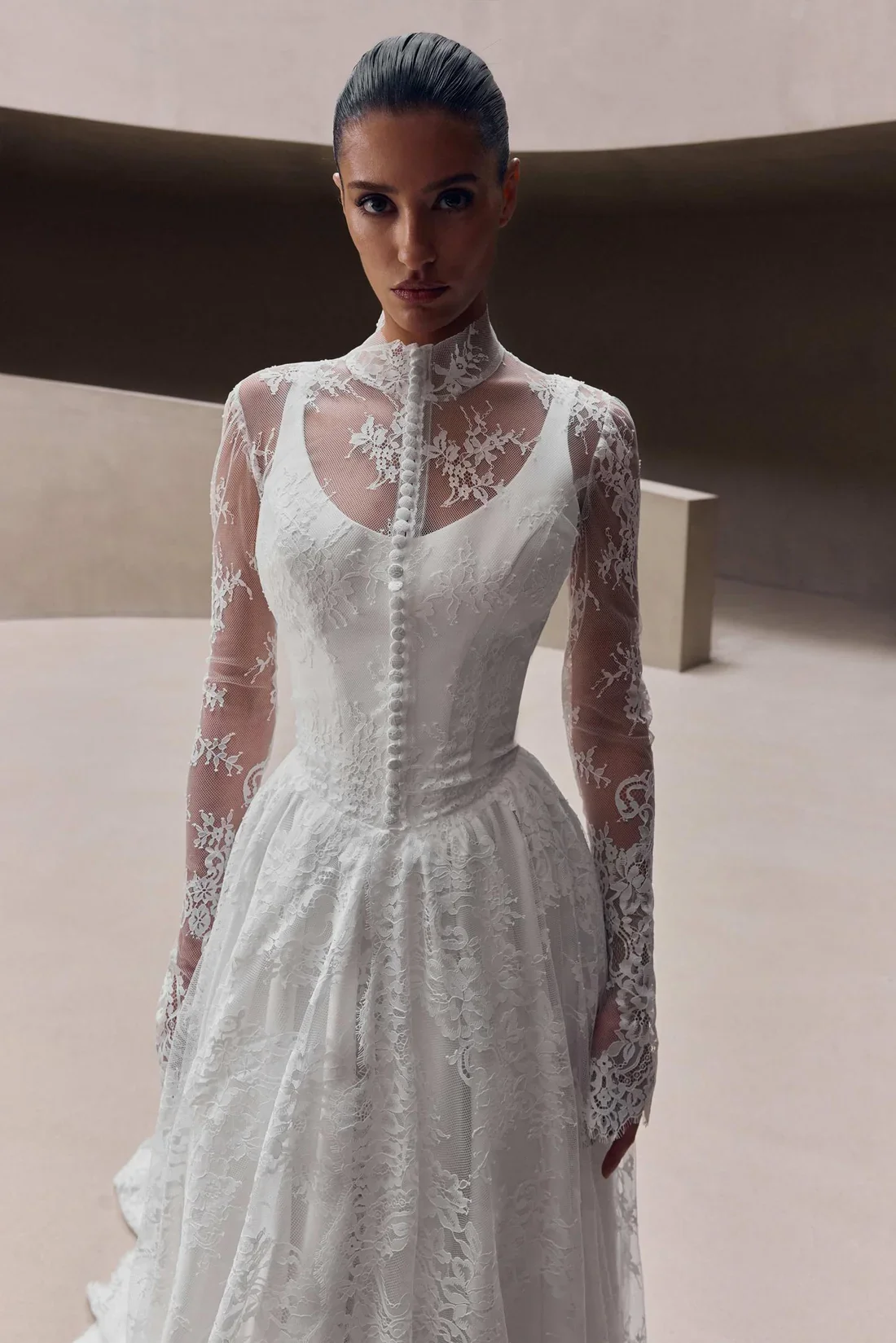 A woman in a white lace wedding dress with long sleeves and button details, standing in front of a modern, minimalist background.