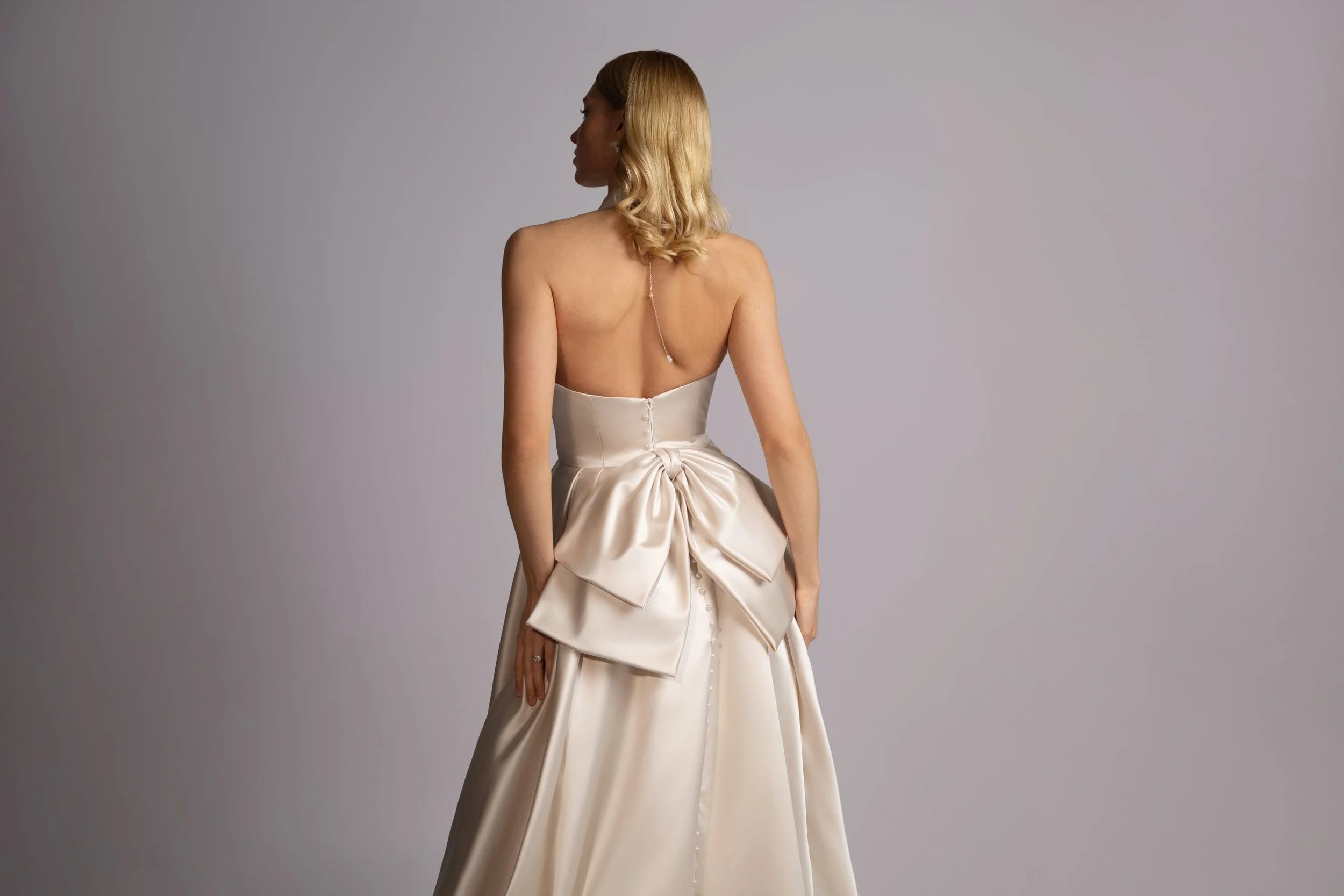 Back view of a woman in a strapless satin wedding gown with large bow and buttons down the back.