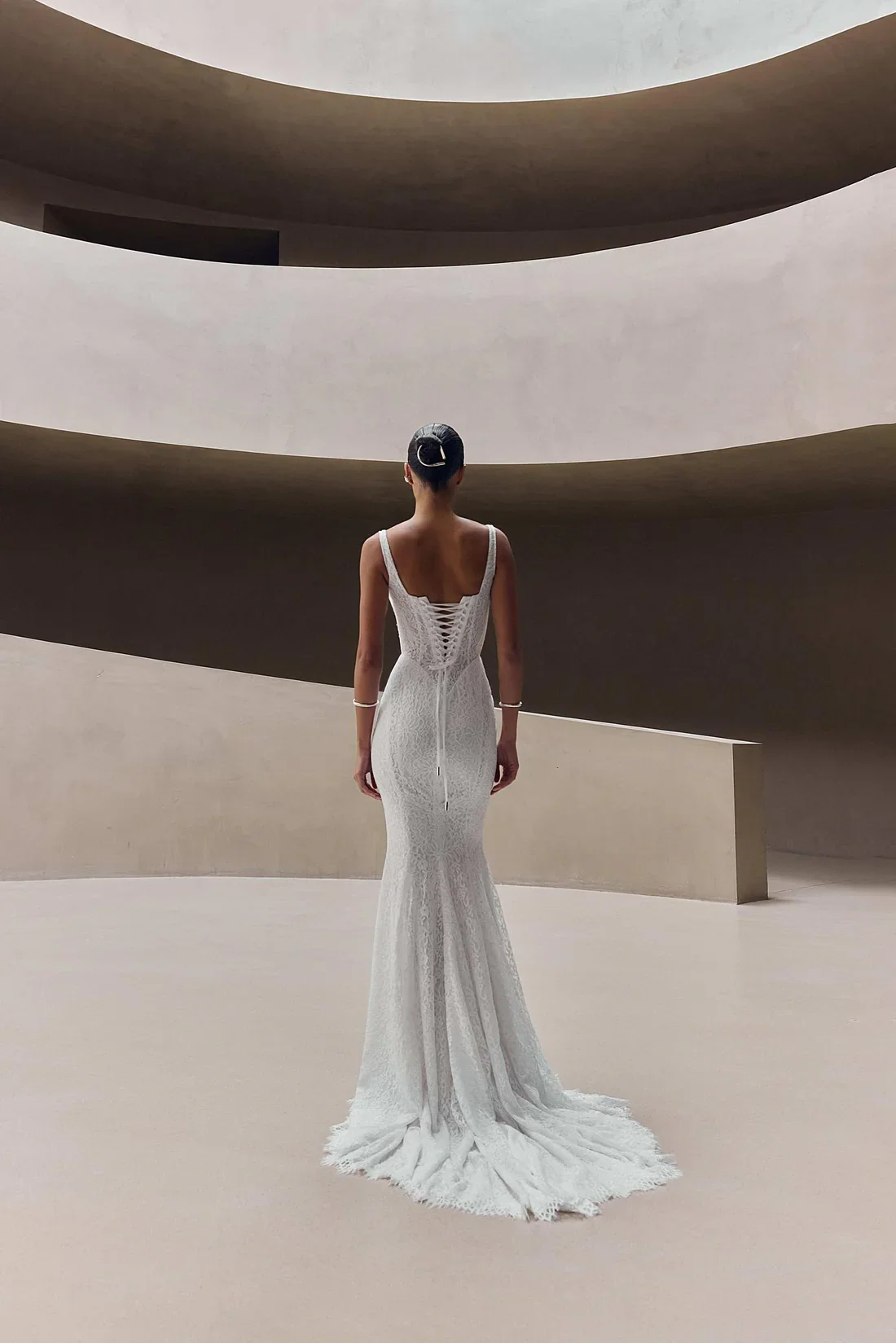 A woman in a white lace wedding dress standing indoors, facing away from the camera, with curved architectural structures in the background.