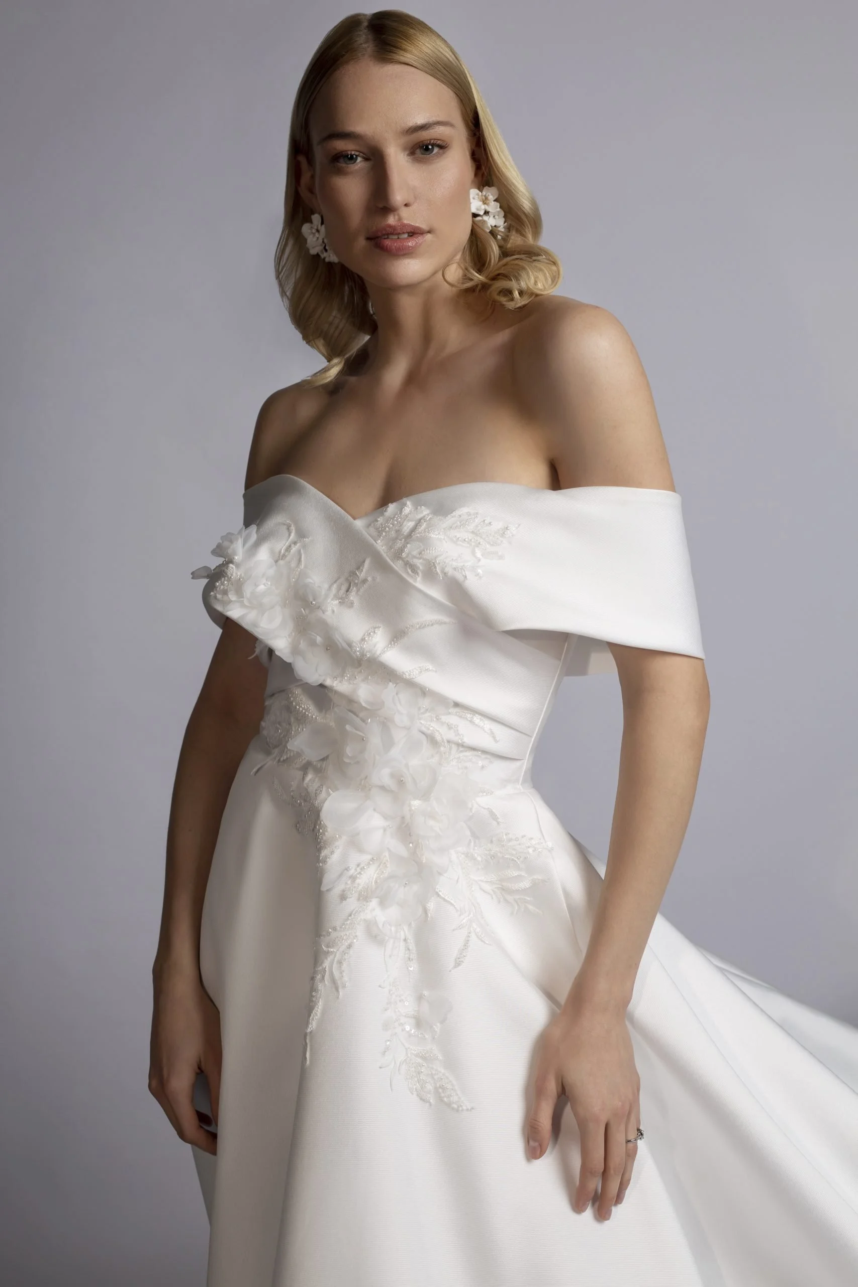 A woman wearing a white off-shoulder wedding dress with floral embroidery and 3D floral appliques, posing against a plain background.