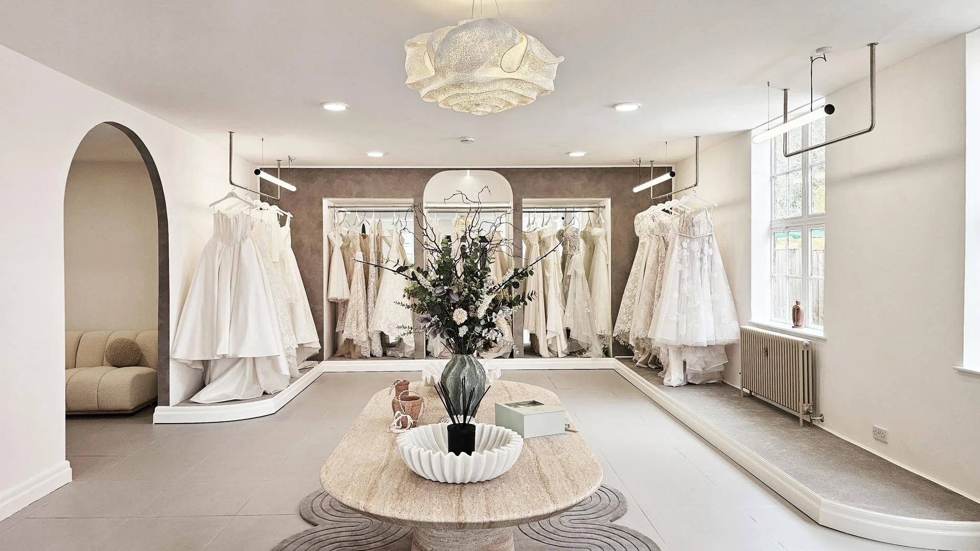 Wedding dress boutique interior with white wedding dresses on display racks, a central floral arrangement on a stone table, large windows with natural light, and minimalistic decor.