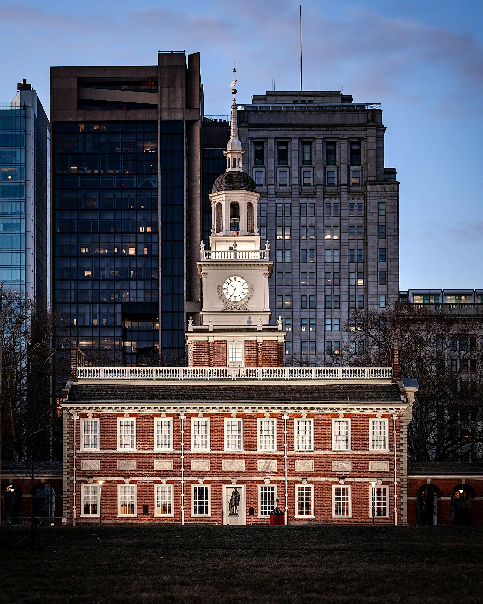 A historic brick building with a clock tower and statue in front, surrounded by modern office buildings, during dusk.